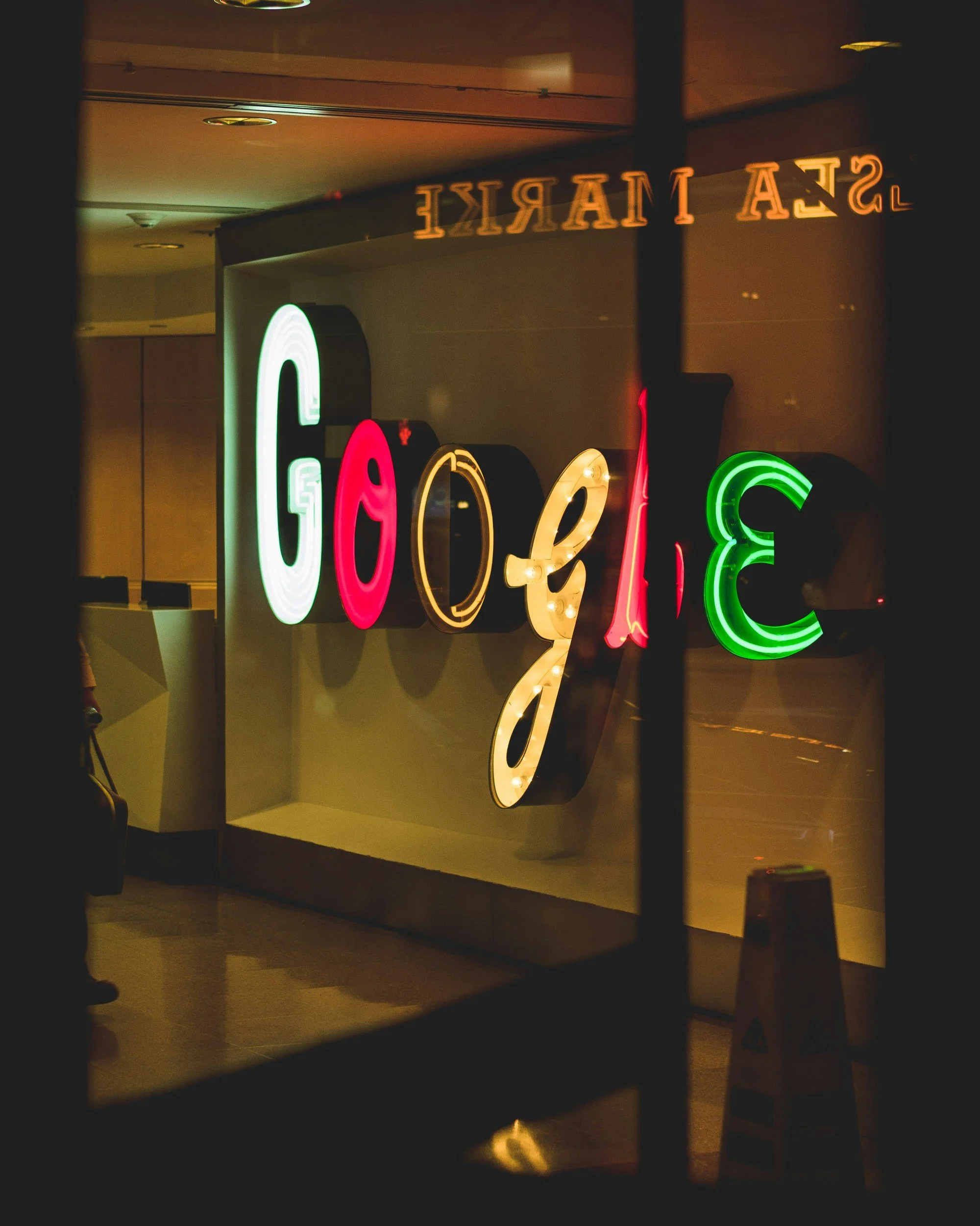 Colorful illuminated sign spelling out "GAY" in various colors, reflected in a mirror. The setting appears to be inside a building with a wooden floor and a wall, and a caution sign is visible in the background.