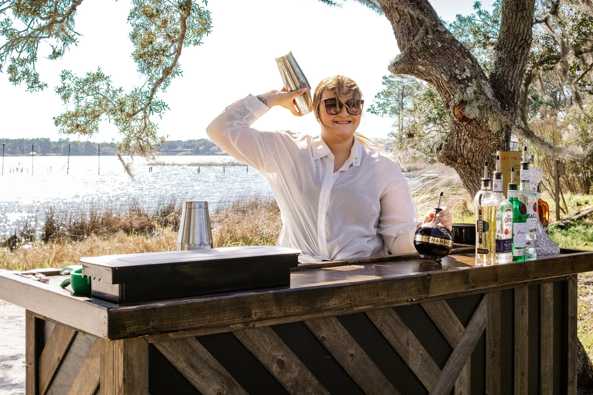 A woman in a white shirt and sunglasses smiling while holding a cocktail shaker at an outdoor bar near a lake, with trees and a bright sky in the background, and bottles of alcohol on the bar table.