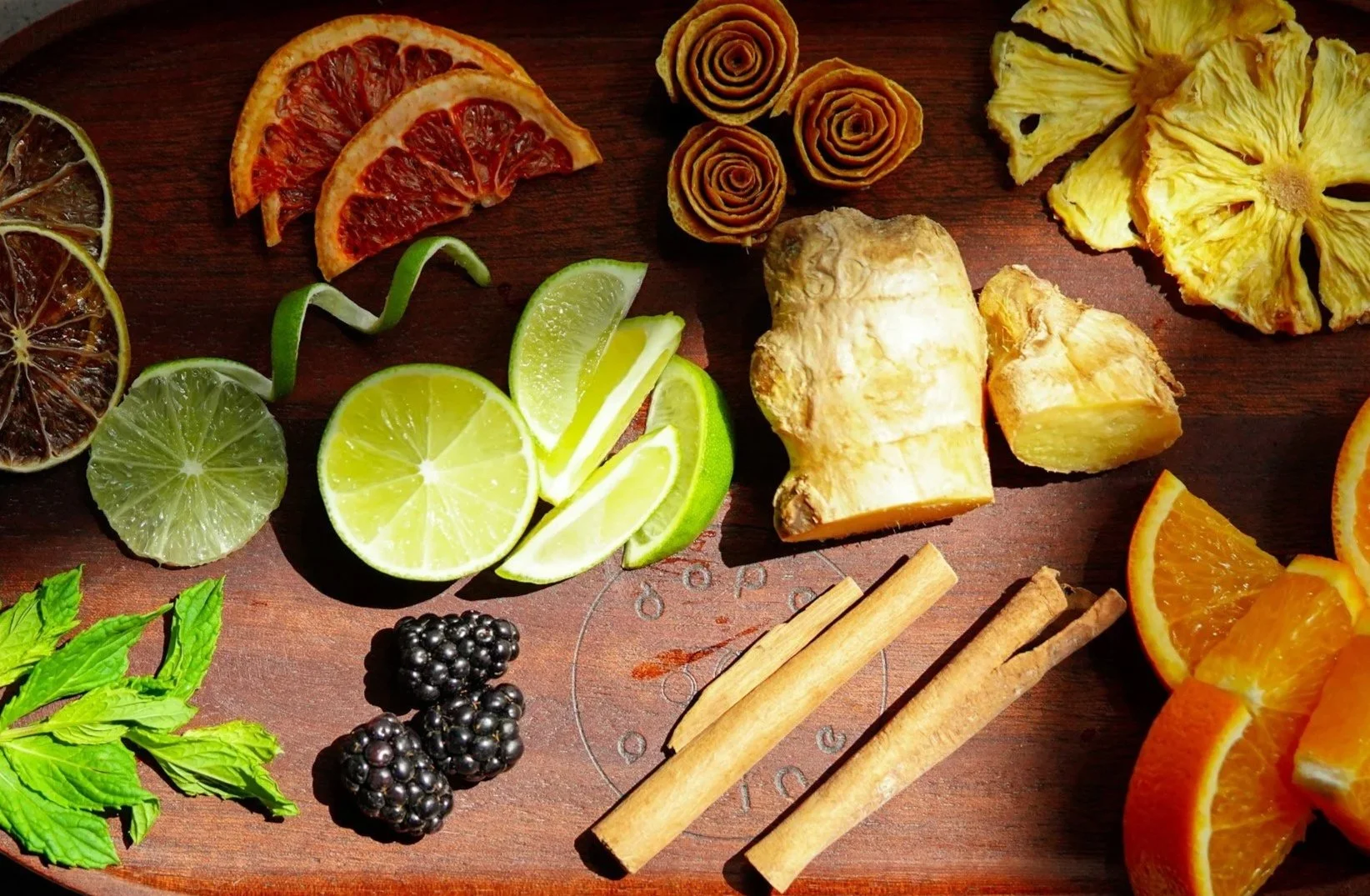Assorted dried and fresh fruits including lemon, lime, orange slices, pineapple, blackberry, and dried citrus slices, with cinnamon sticks and fresh mint leaves on a wooden surface.