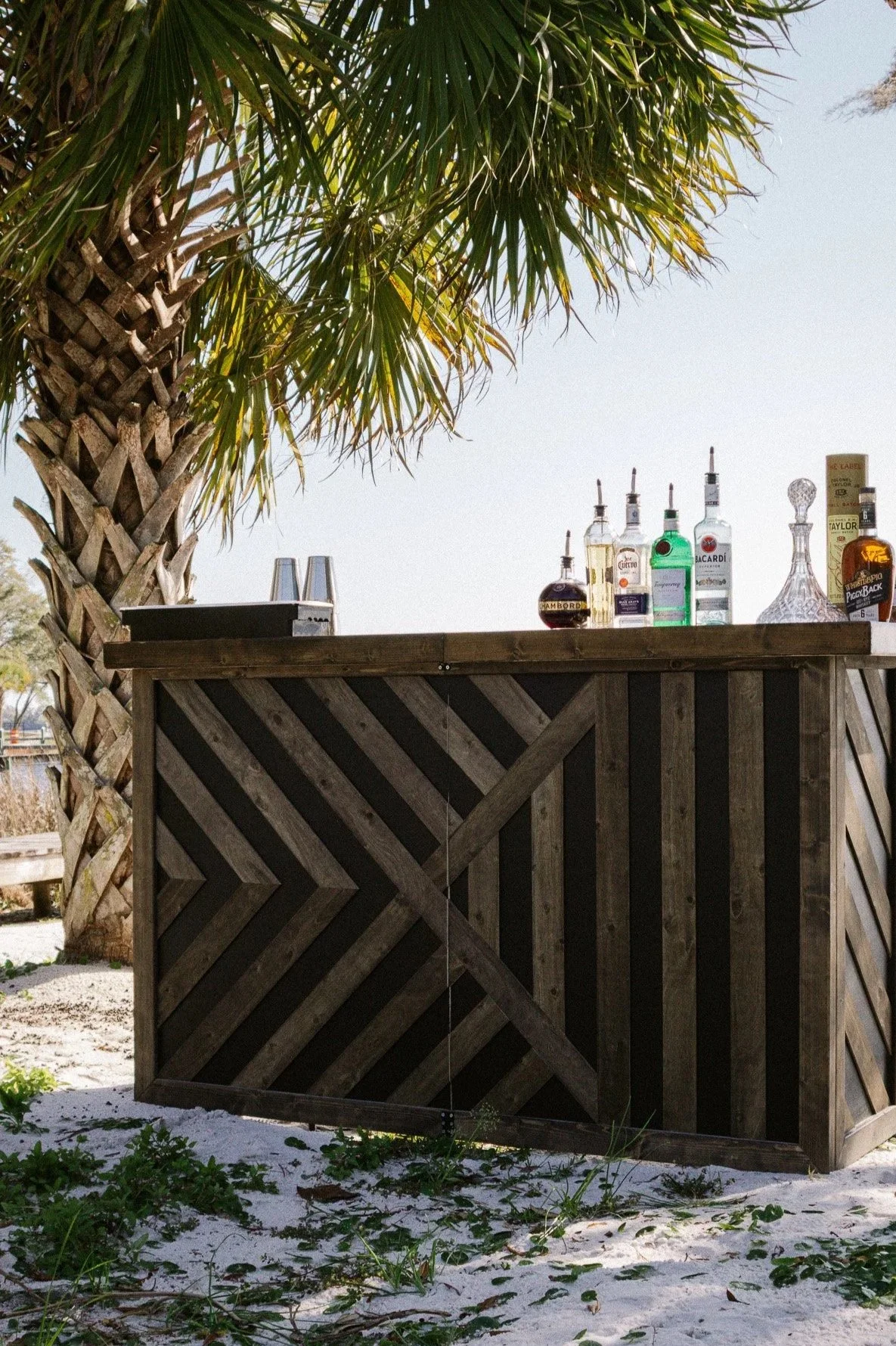 A wooden bar with various alcohol bottles and two empty glasses, set in a tropical outdoor location with a palm tree overhead.
