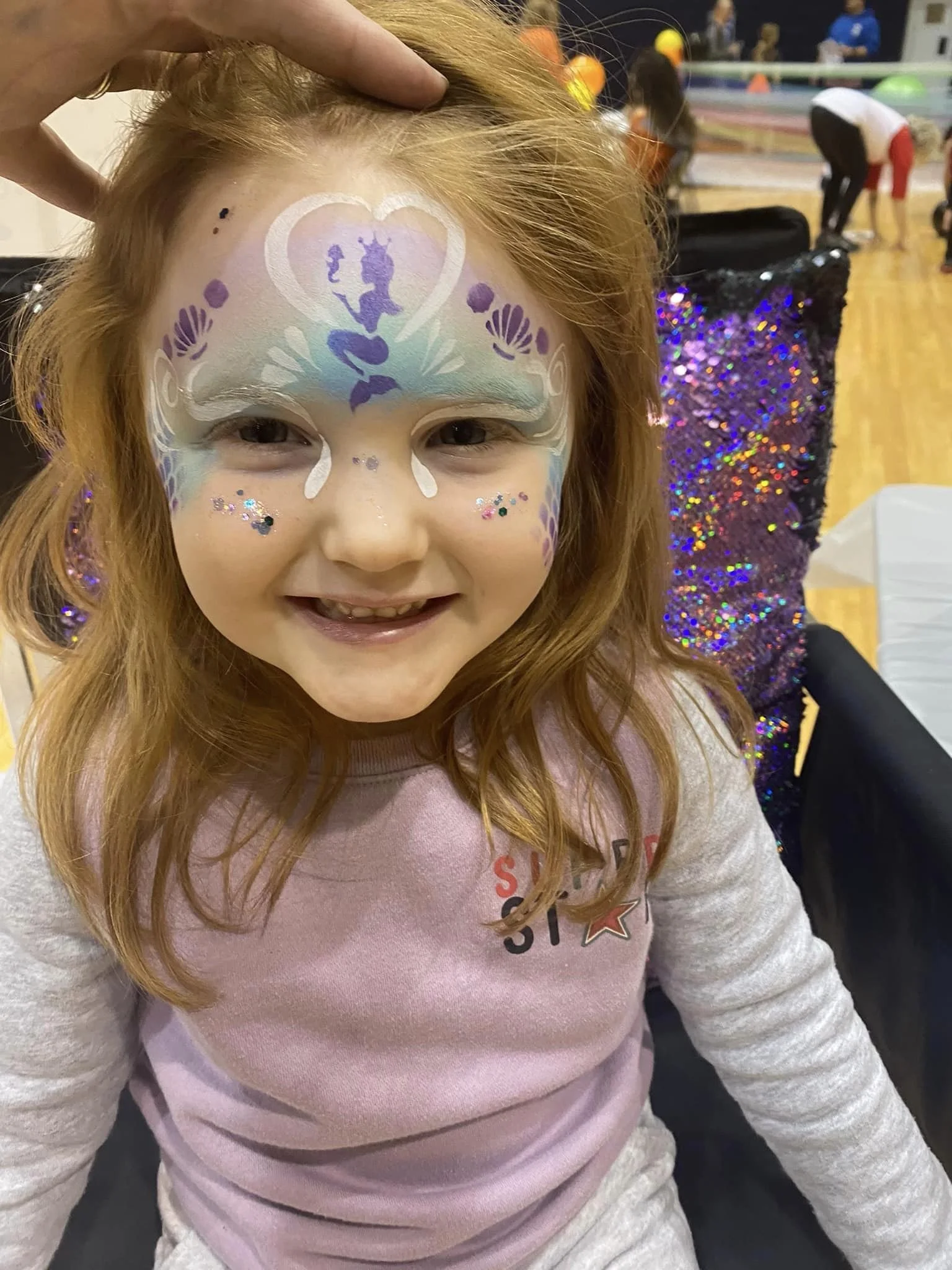 A young girl with red hair has a colorful fairy face paint design, featuring a butterfly, hearts, and floral details, with glitter and sequins on her cheeks, at an indoor event with people in the background.
