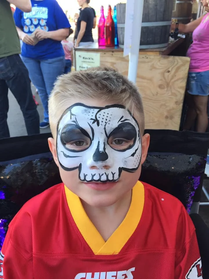 A young boy with half of his face painted as a skull, smiling at the camera. He's wearing a red sports jersey with yellow accents, at an outdoor event with people and colorful bottles in the background.
