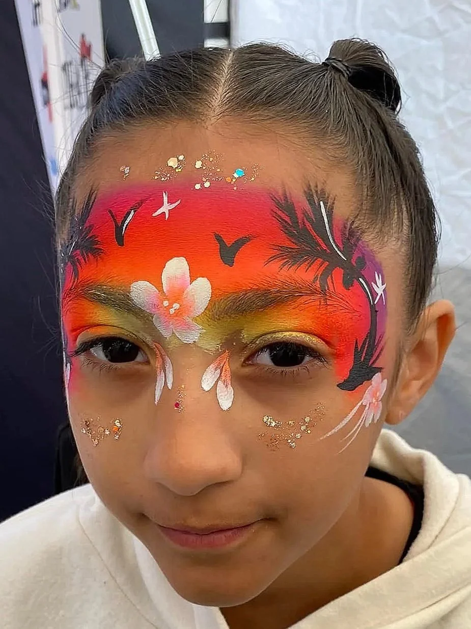 A young girl with hair styled in two buns has elaborate face paint featuring a flaming sunset, black silhouettes of birds, pink flowers, and glitter accents on her face.