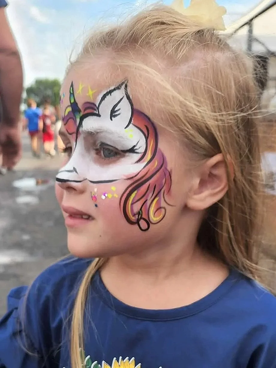 A young girl with blonde hair and face paint of a unicorn, with rainbow colors at an outdoor event with people and a wooden stand in the background.