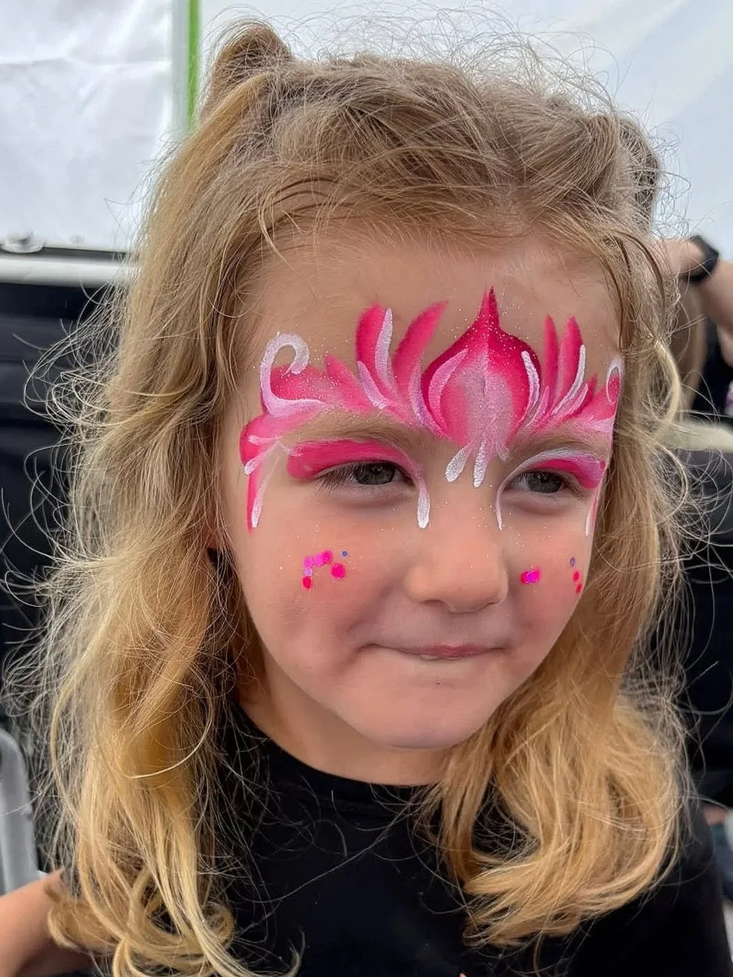 A young girl with curly blonde hair and face paint designs of a pink glittery crown. 