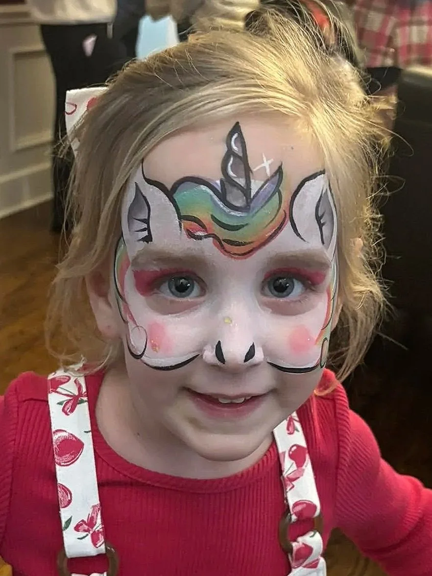 A young girl with blonde hair and blue eyes, smiling, has face paint resembling a unicorn with a rainbow mane and ears. 