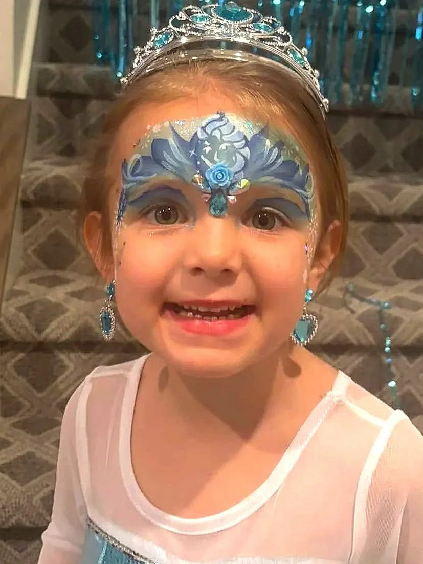 A young girl dressed as a princess, wearing a silver tiara and an Elsa costume from Frozen, standing at the bottom of a staircase decorated with blue metallic streamers.