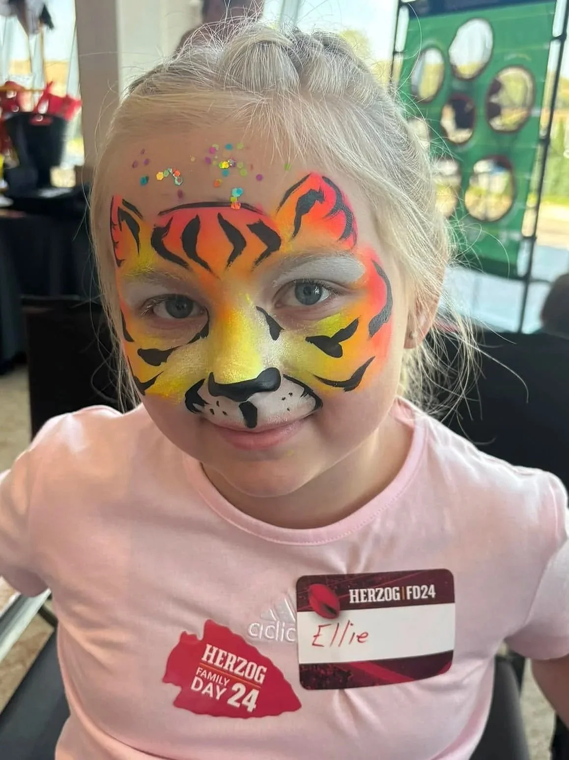 A young girl with face paint resembling a tiger, featuring orange, black, and yellow colors with confetti on her forehead.  Herszog Family Day 24 at the Chiefs Stadium. 