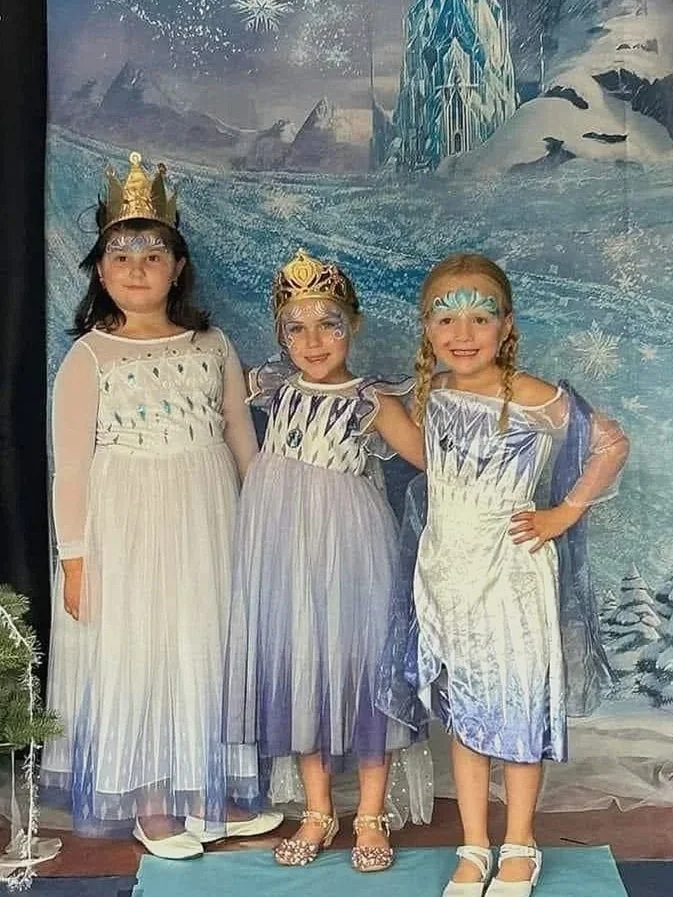 Three young girls dressed as princesses or ice fairies, standing in front of a winter or ice kingdom-themed backdrop, at the GEHS theatre frozen production.