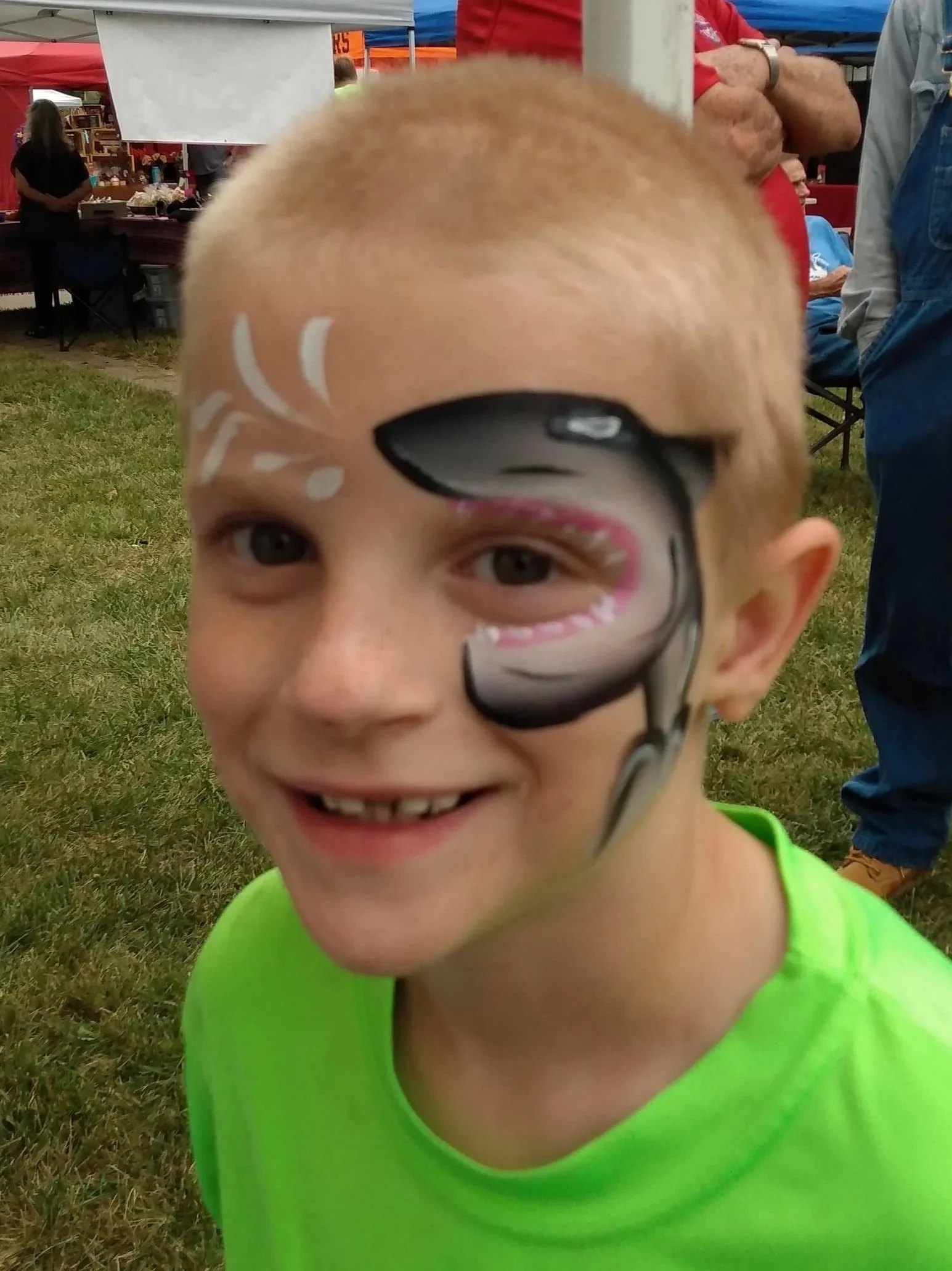 A young boy with face paint smiling at the camera at an outdoor event.