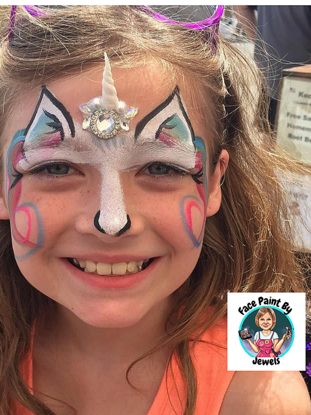 A smiling young girl with a unicorn face paint design, including a white horn with a jewel, white and black detailing, colorful accents around her eyes, and a nose line.