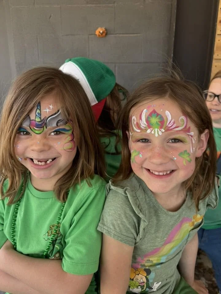 Two young girls with face paint smiling, dressed in green shirts, celebrating St. Patrick's Day.
