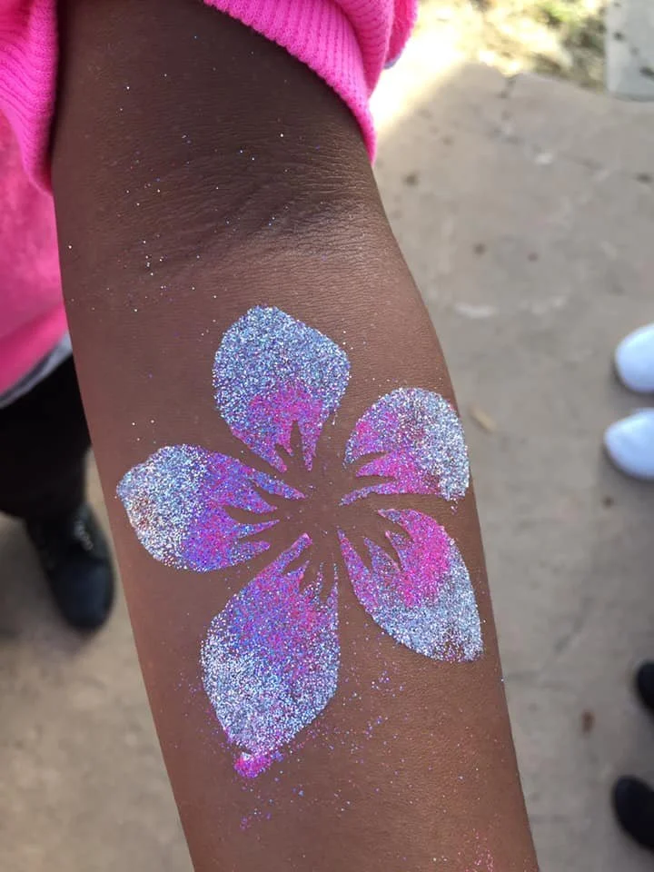 A glittery flower design on a person's arm with five petals in white, pink, and purple hues, on tan skin, with a pink sleeve partially visible.