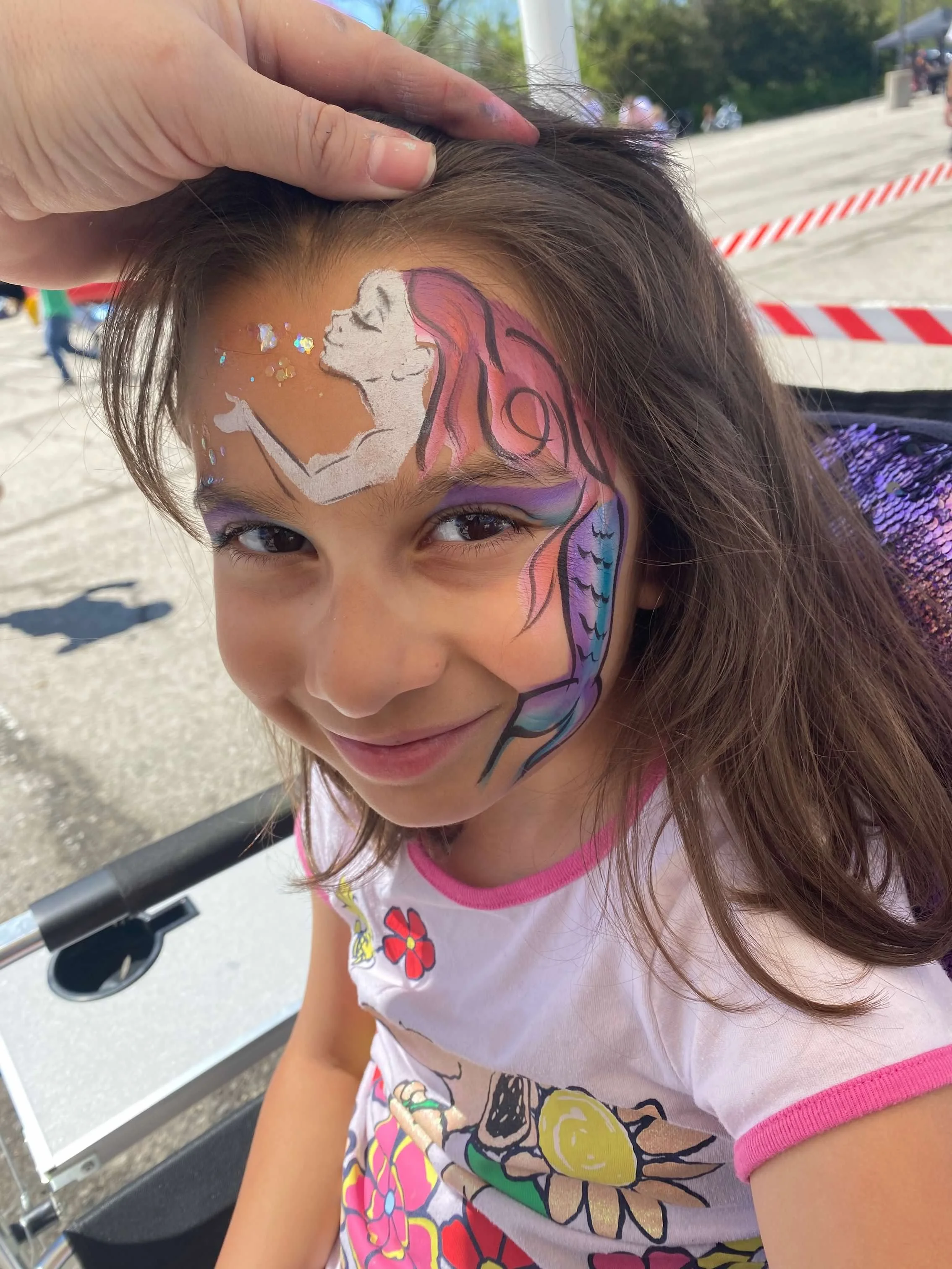Girl with face paint of a colorful mermaid with pink, purple, and blue shades, and a white unicorn on her forehead, at an outdoor event.