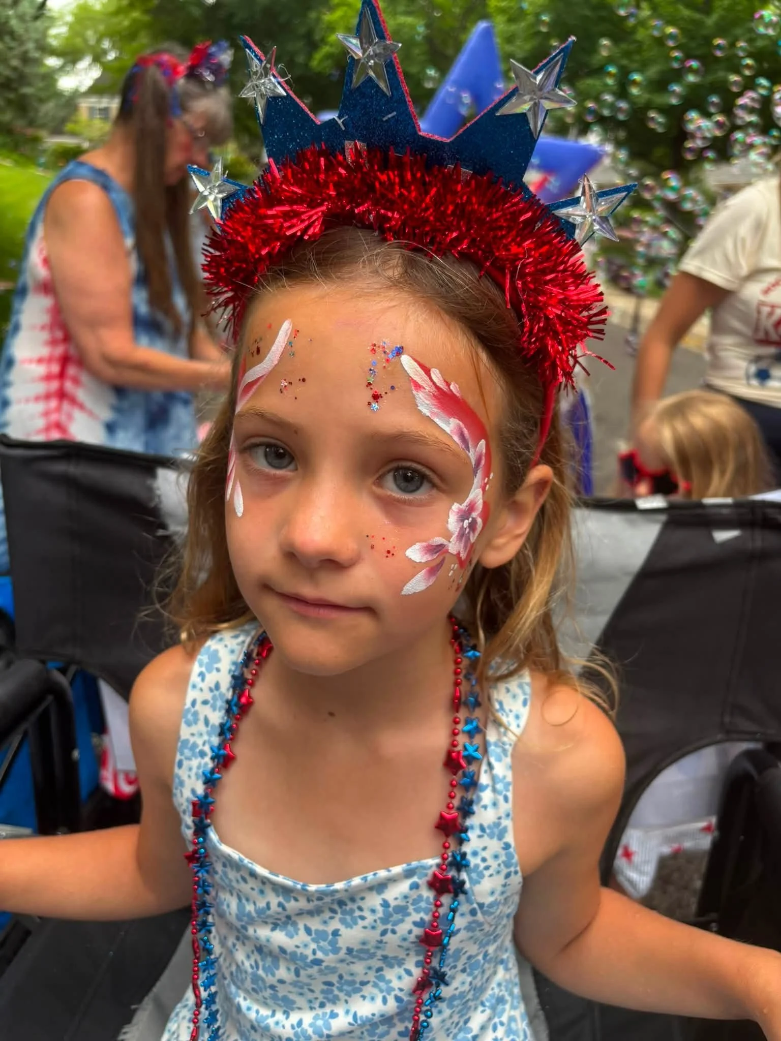 Young girl with face paint and patriotic decorations wearing a headband and necklaces, celebrating the 4th of July.