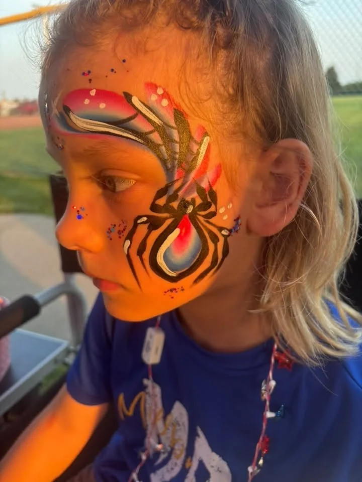 Close-up of a young girl with face paint resembling a spider on her cheek, with a background of a sports field during sunset.