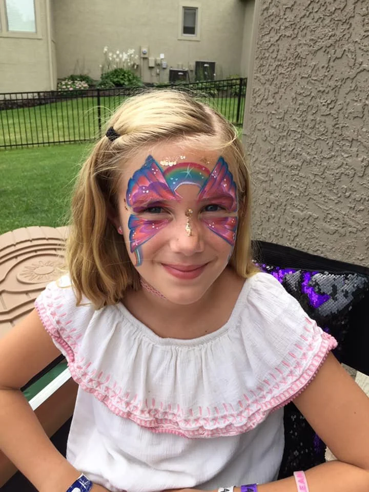 A young girl with butterfly face paint featuring a rainbow and stars, smiling outdoors at her birthday party.