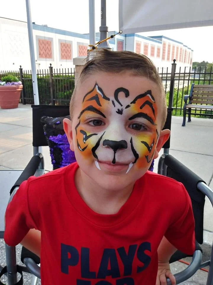 A young boy with face paint resembling a tiger, sitting in a wheelchair outdoors, wearing a red shirt that says 'PLAYS' in blue letters, under a white canopy with a fence and building in the background.