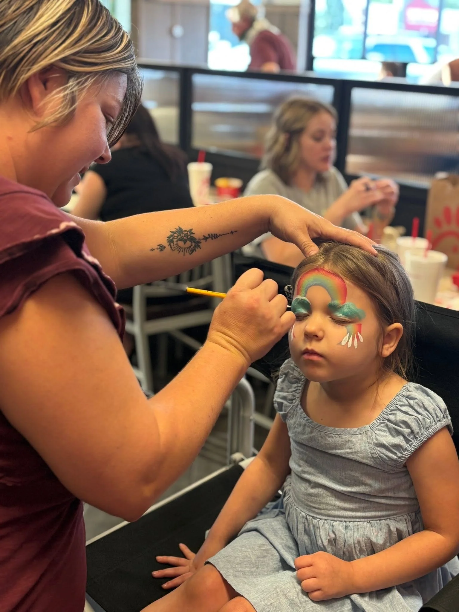 A young girl with face paint of a rainbow and clouds is sitting with her eyes closed at a local chick-fil-a event. 
