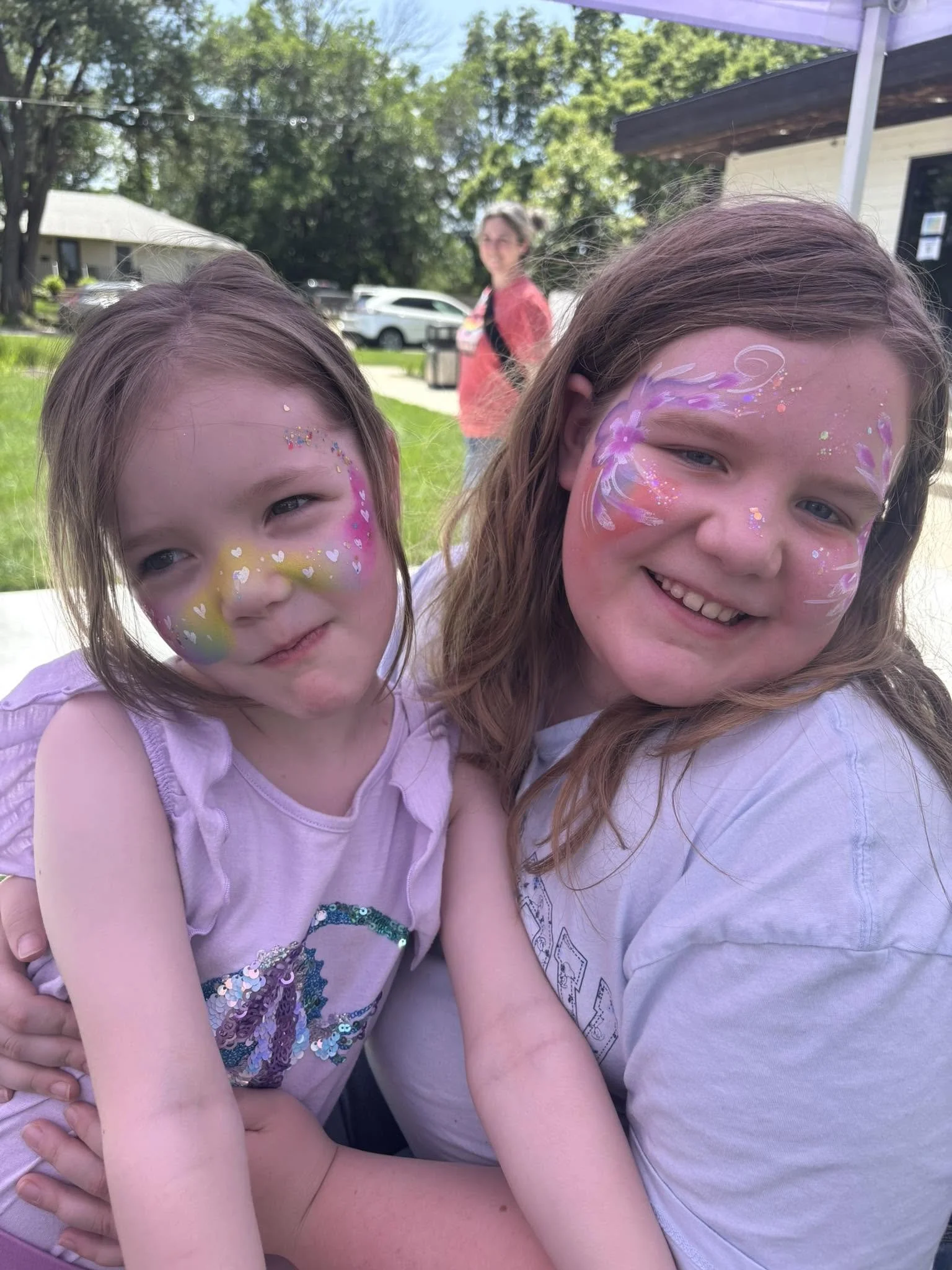 Two young girls with colorful face paint, smiling and sitting close together at a festival.