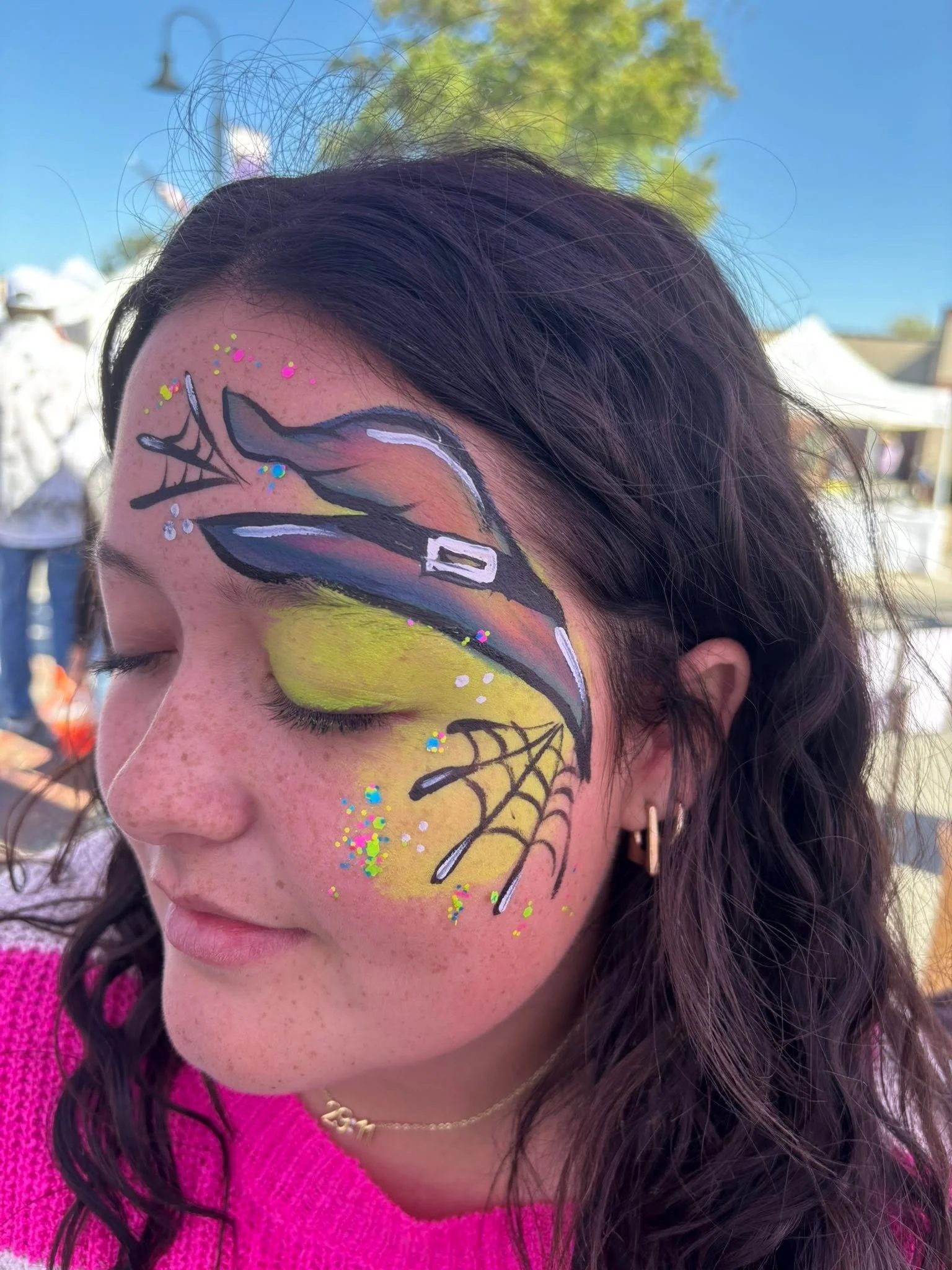 Close-up of a woman's face with face paint featuring a colorful witches hat, glitter, and decorative accents, wearing a pink sweater and black earrings, outdoors on a sunny day.