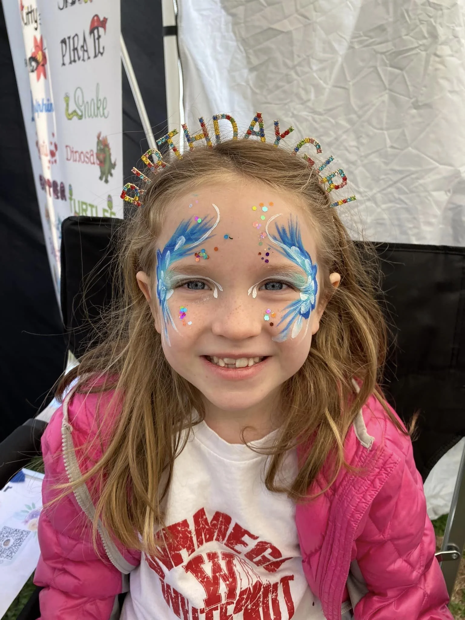 Young girl with face paint of blue and white waves, colorful glitter, and white accents, wearing a birthday tiara that spells out 'HAPPY BIRTHDAY', pink jacket, and white T-shirt with red text.