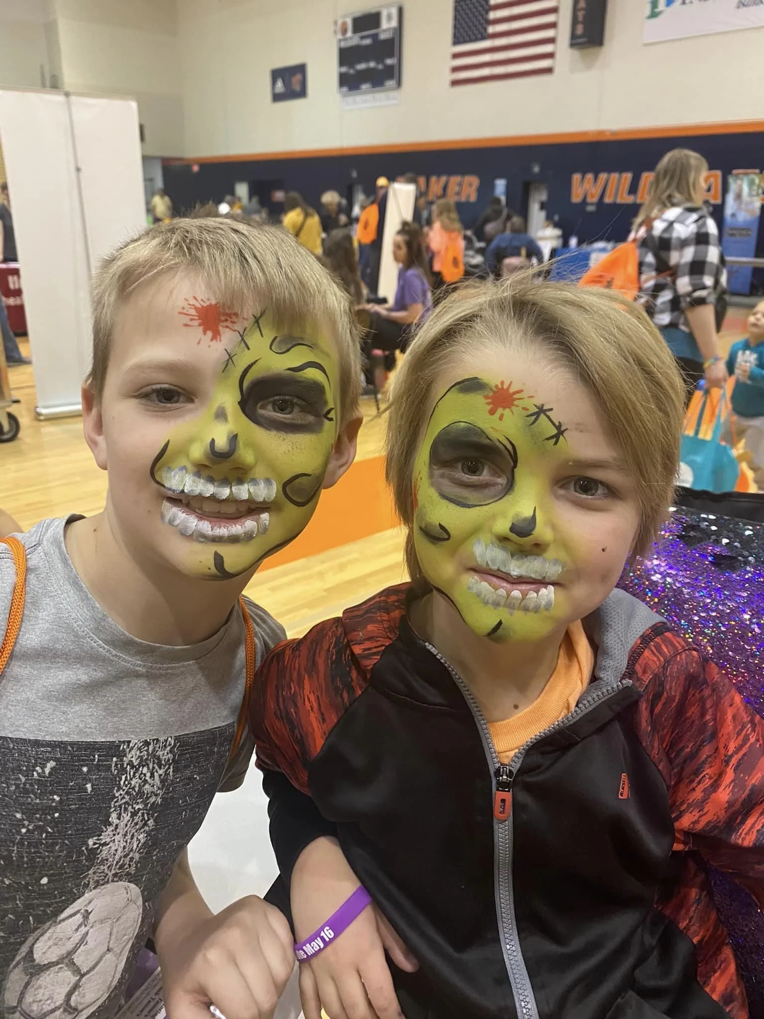 Two children with face paint resembling skeletons, smiling at a school event in a gymnasium.