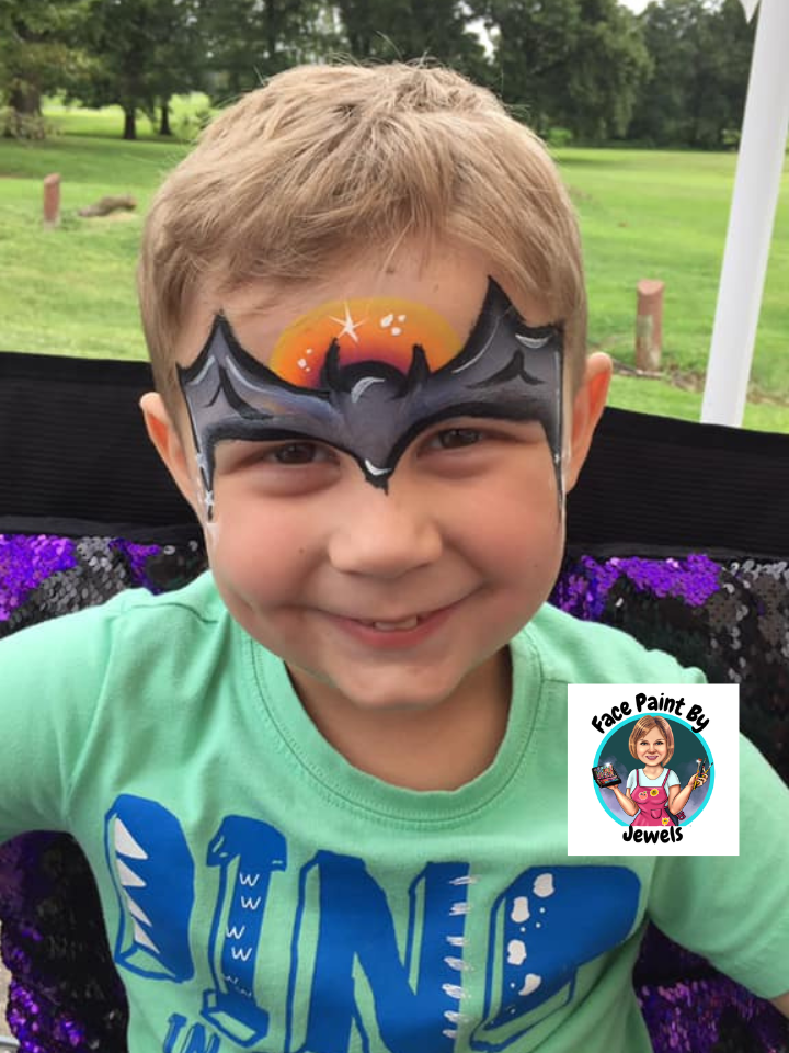 A young boy with a face paint of a bat symbol on his forehead, smiling outdoors with green grass and trees in the background.