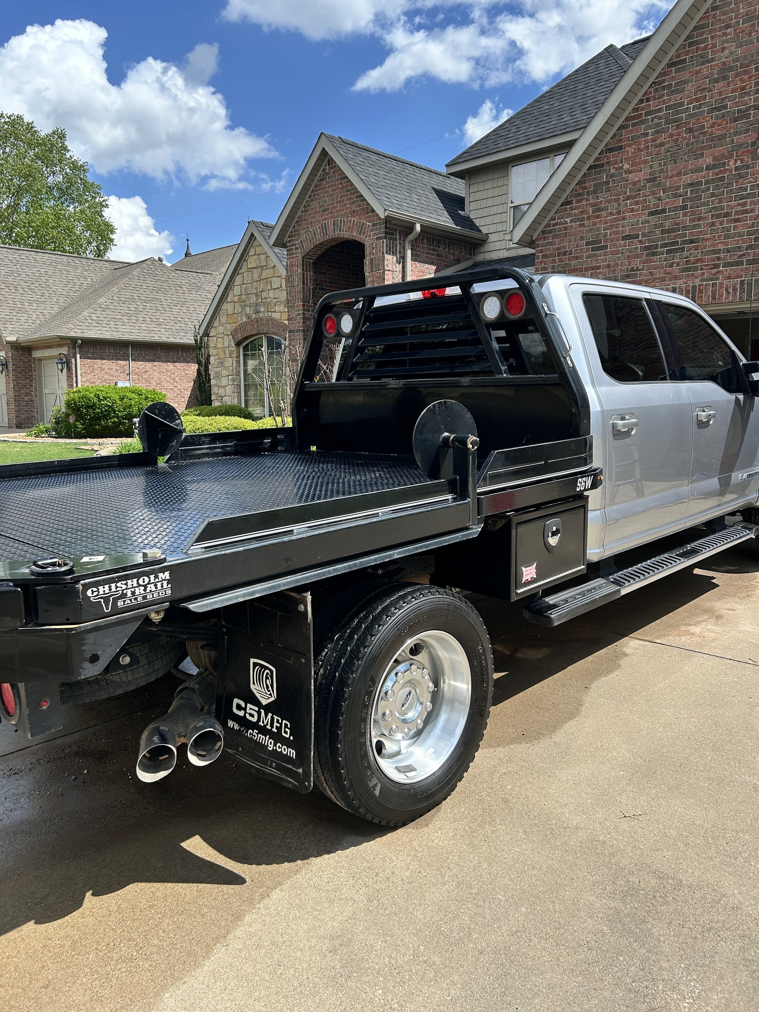 Silver pickup truck with a flatbed parked on a residential driveway in front of brick and stone houses, under a partly cloudy sky.