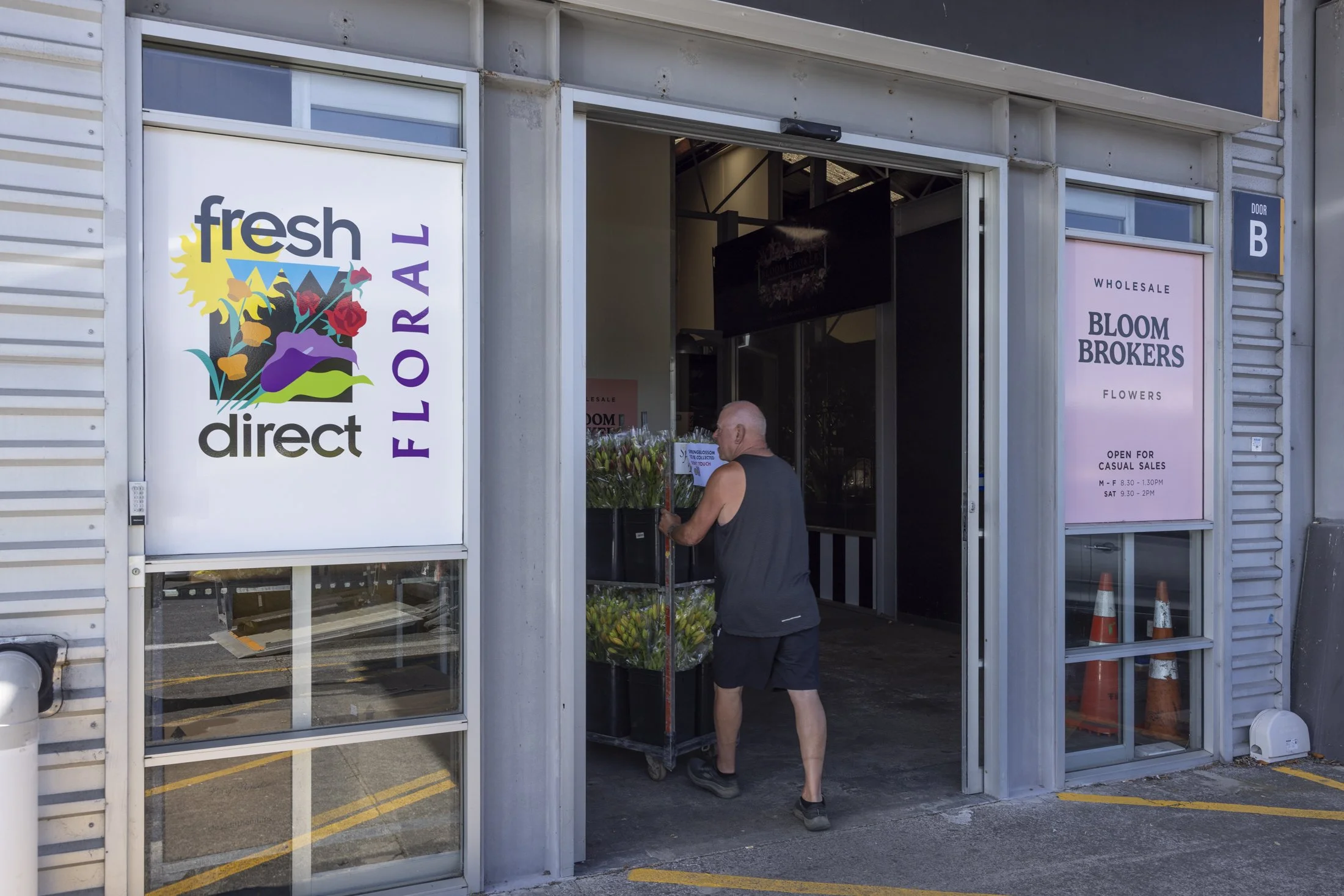 A man shopping for flowers at a florist, entering a store called 'Fresh Direct Floral' with signs for 'Bloom Brokers' and casual sales hours.