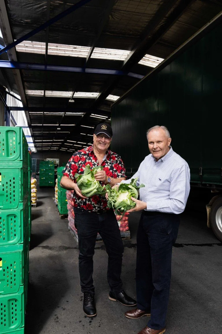 Two men inside a warehouse or market area holding cabbages, smiling at the camera. One is wearing a floral shirt and cap, the other a light-colored shirt. Green crates and a truck are visible in the background.