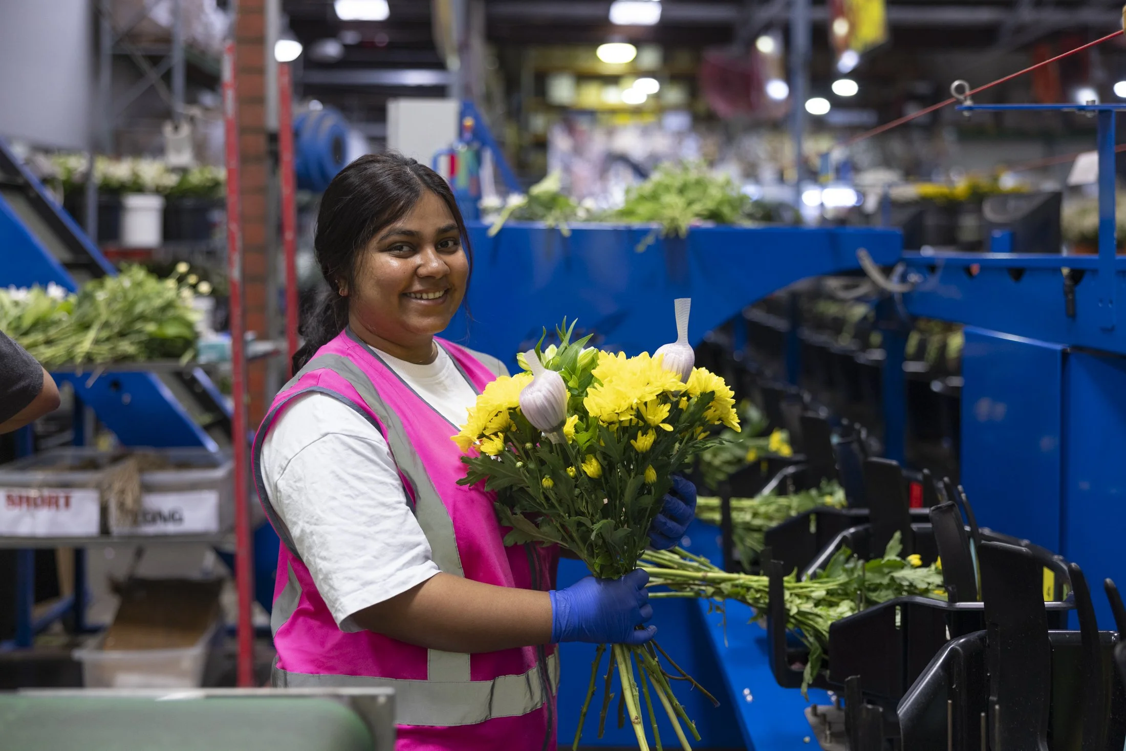 A woman in a pink vest and blue gloves holding a bouquet of yellow flowers with garlic bulbs, smiling in a floral warehouse.