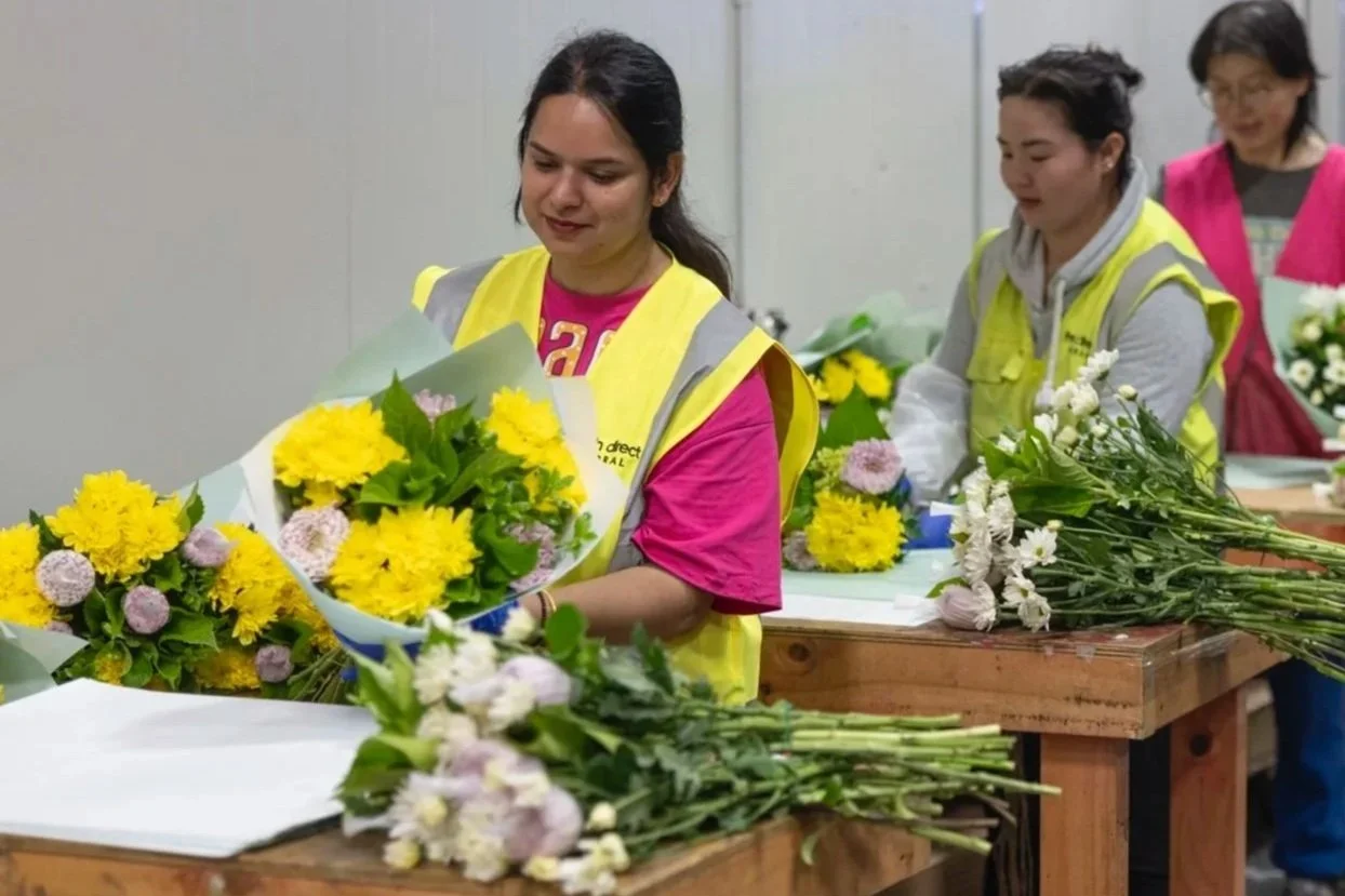 Women in safety vests arranging and wrapping yellow and white flowers on a table in a floral workshop.