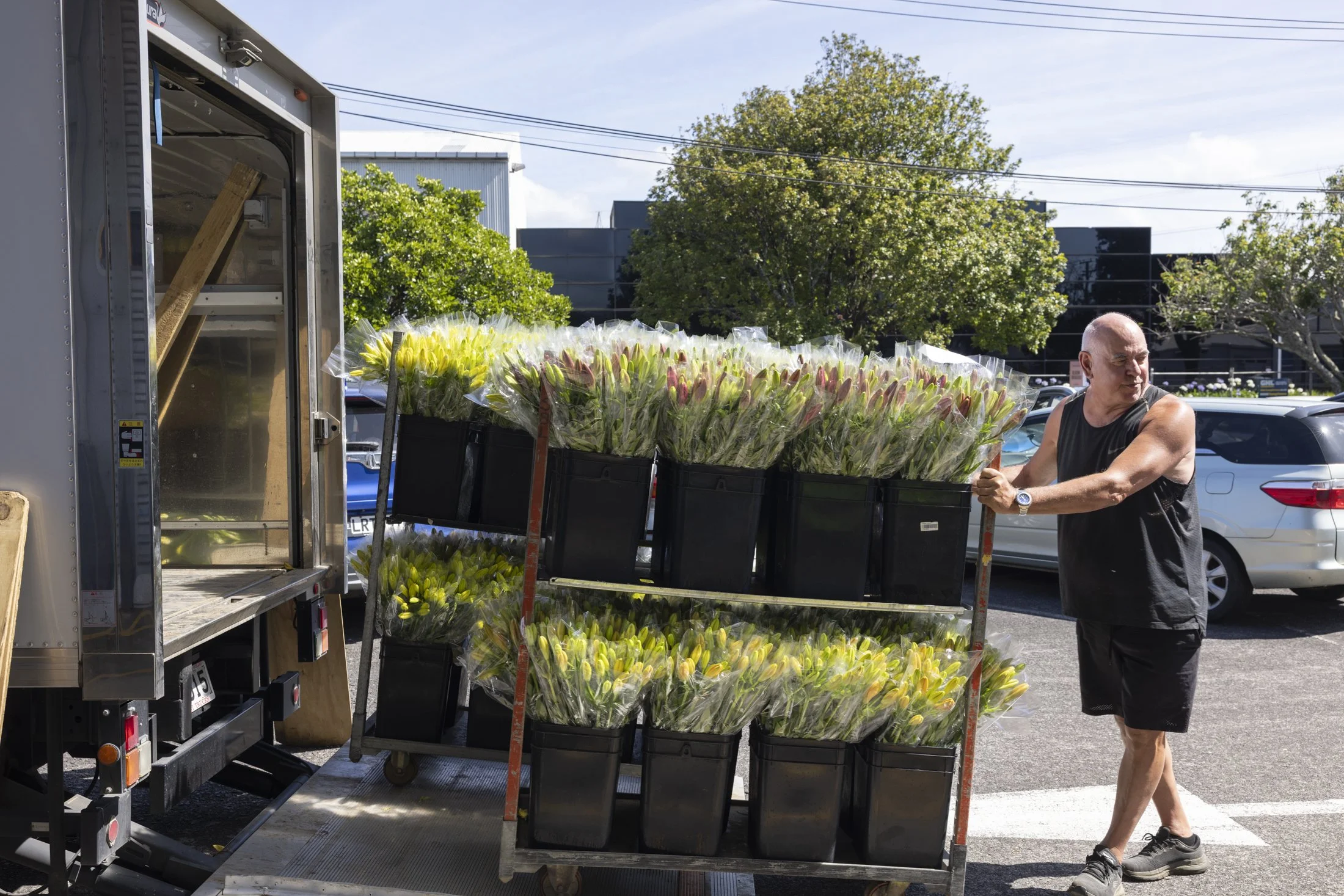 A man in a black sleeveless shirt and shorts pushes a cart filled with bouquets of yellow and pink flowers, parked next to a truck in a parking lot with trees and a building in the background.
