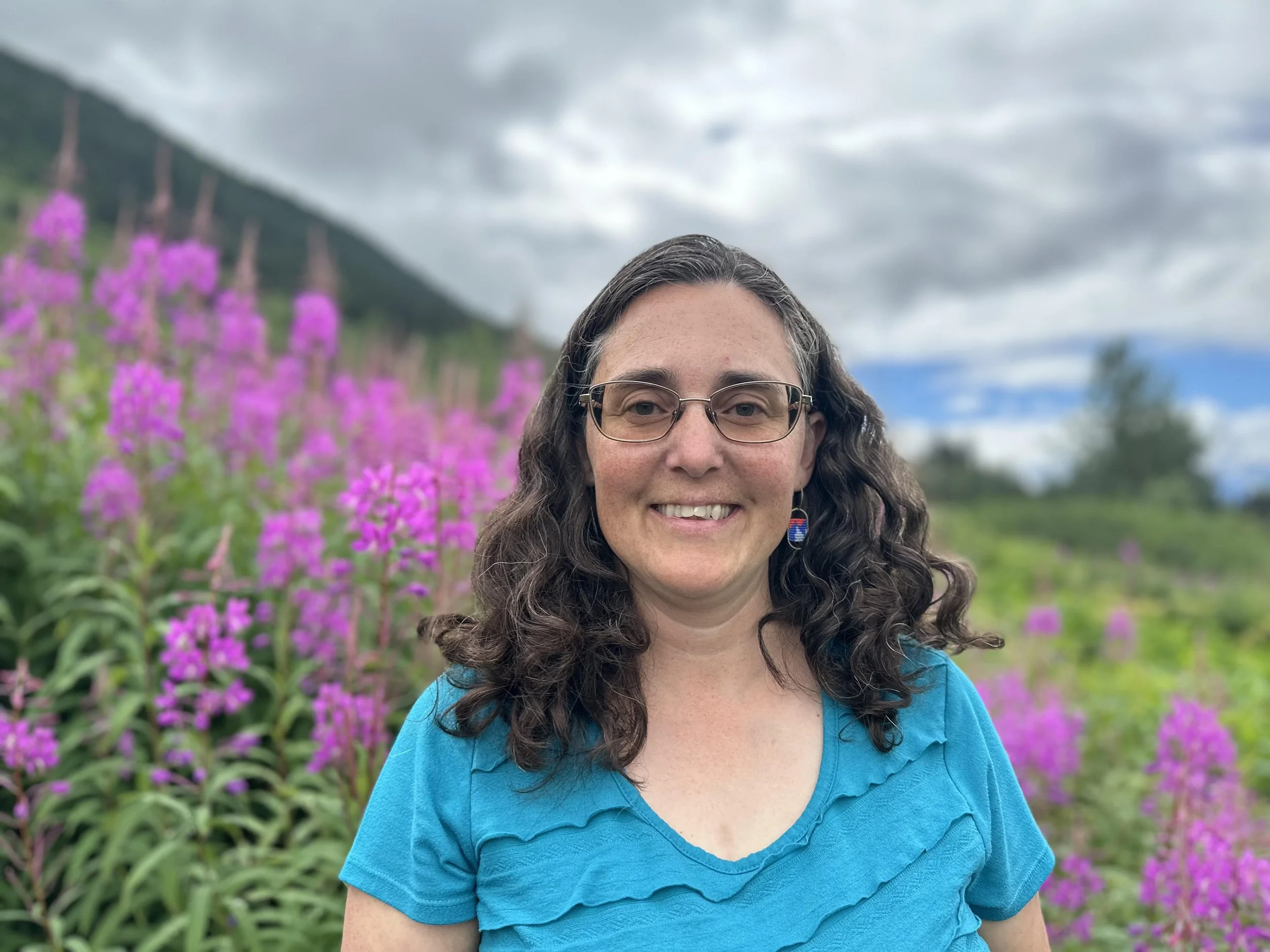 Melissa with glasses and long curly hair smiling outdoors in a field of pink flowers with mountains and cloudy sky in the background.