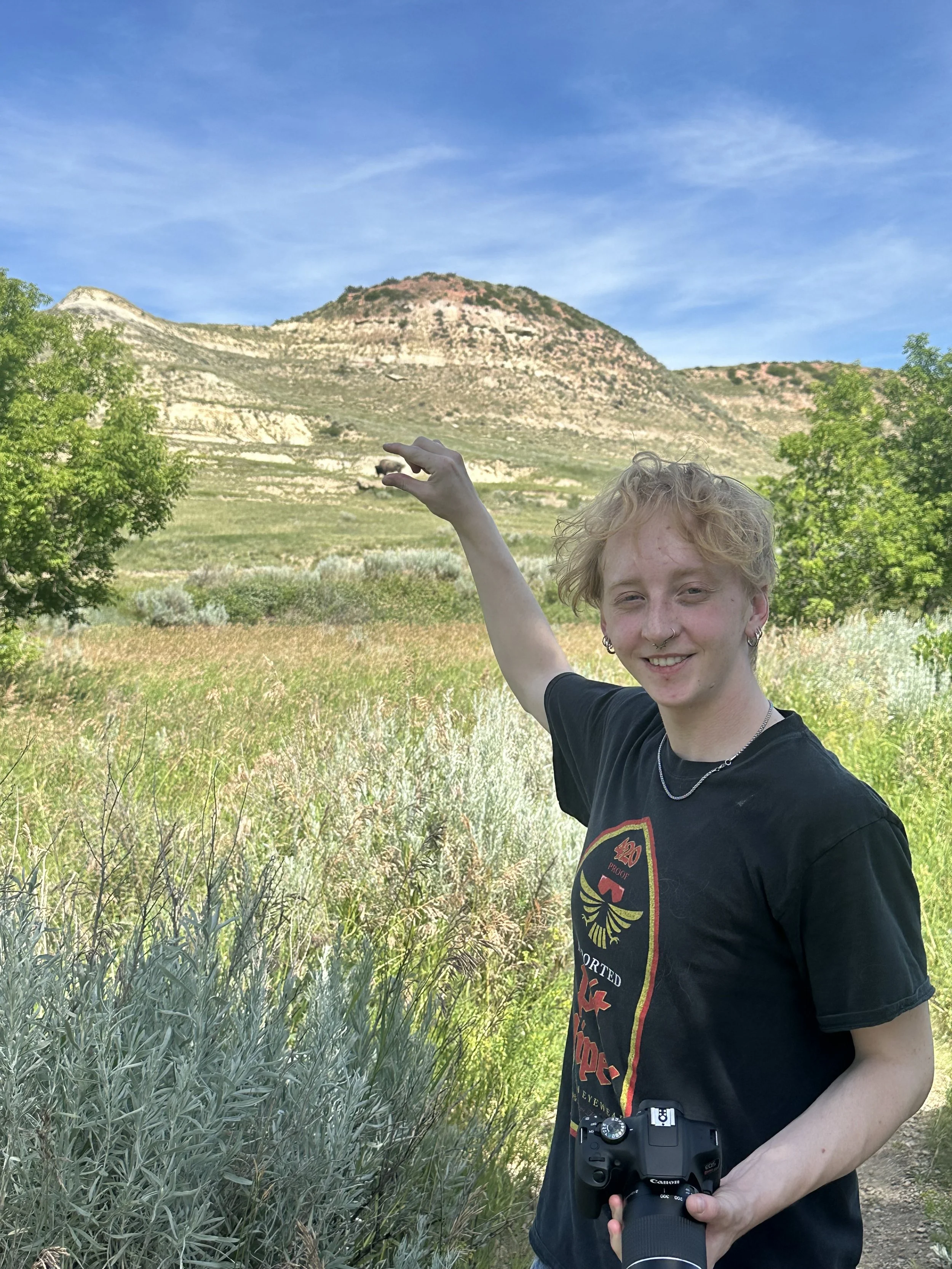 A young person with curly blonde hair smiling, holding a camera, standing in a field with trees and tall grass, pointing towards a mountain in the background under a blue sky.