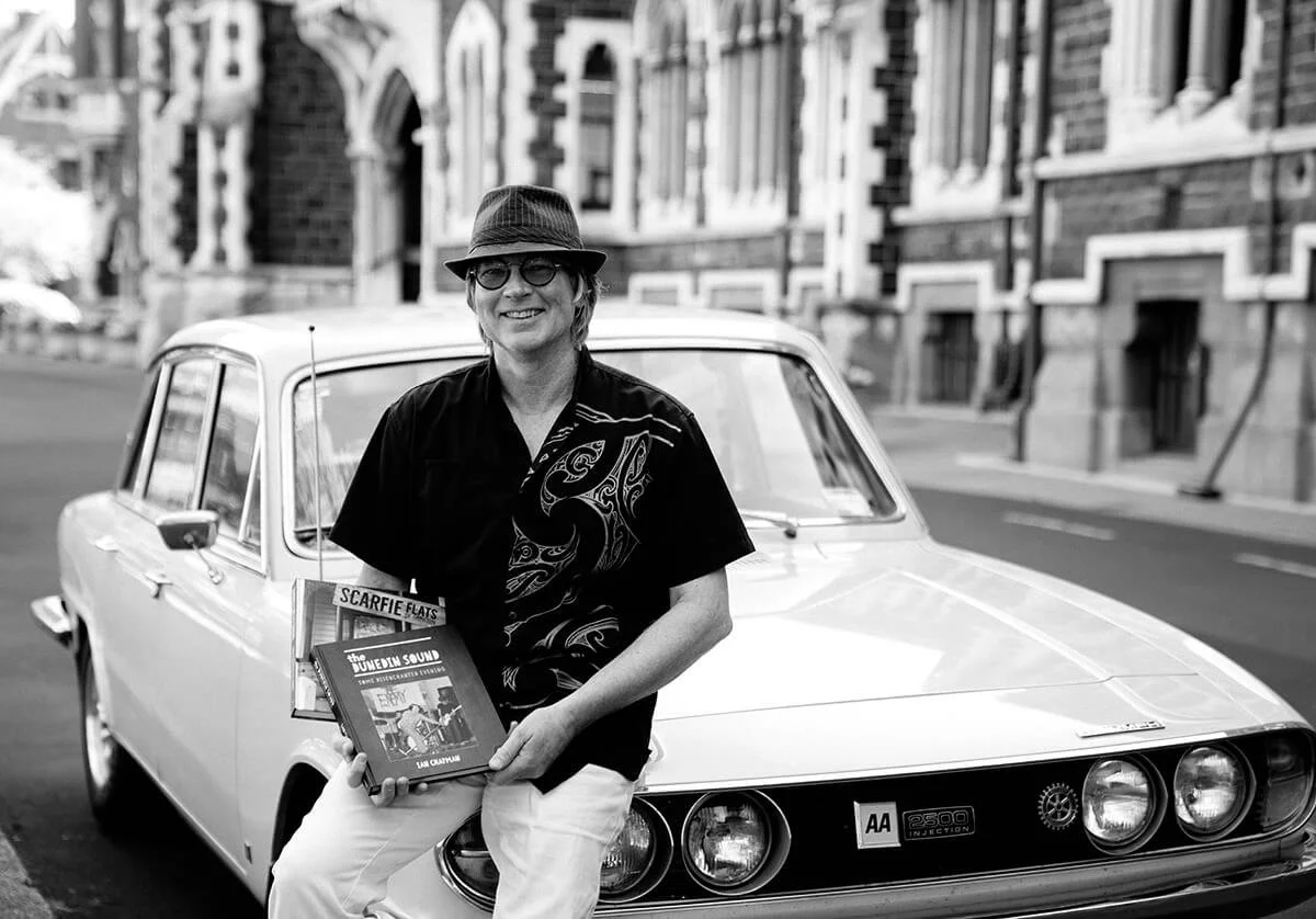 A person sitting on the hood of a vintage car, holding books, on a city street.