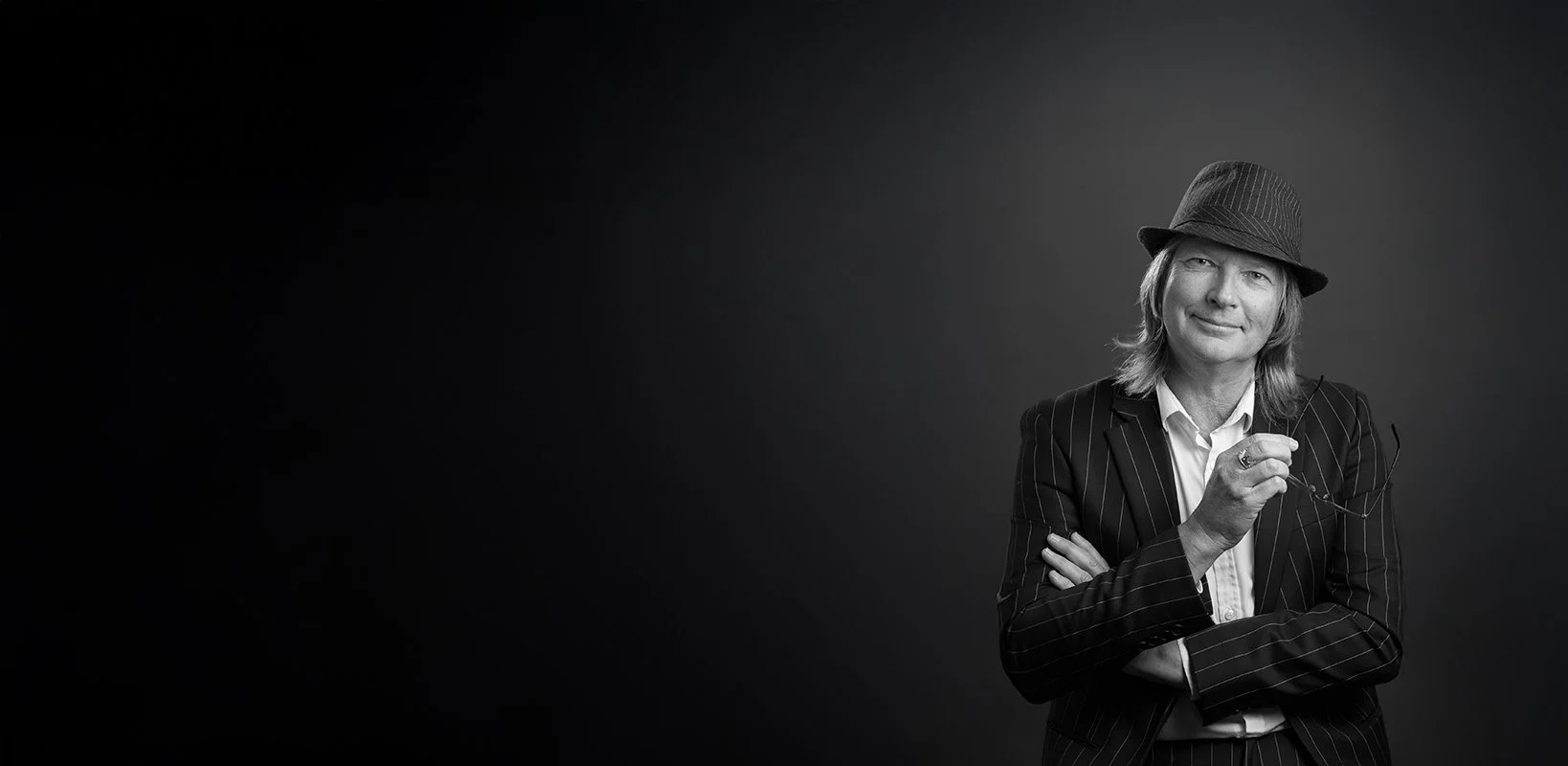 A black and white portrait of a man wearing a pinstripe suit, a white shirt, and a fedora hat. He is holding a pair of eyeglasses and has a slight smile with arms crossed.