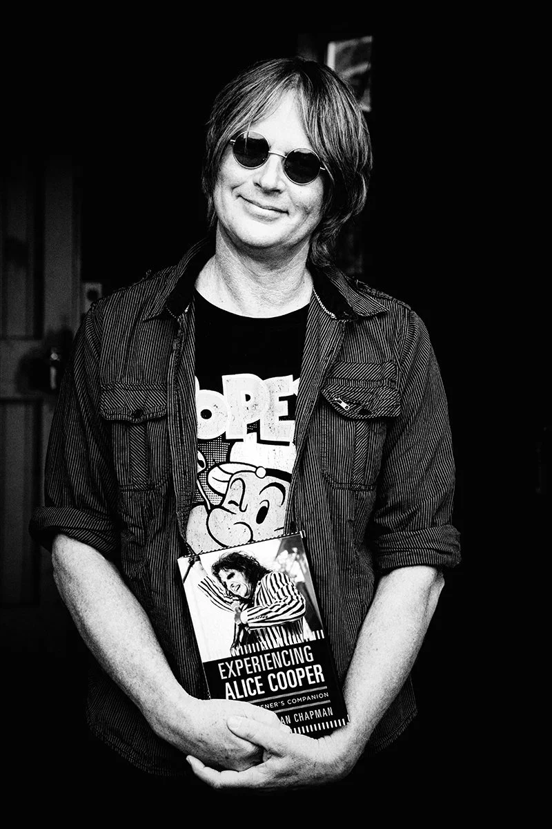 A person with shoulder-length hair wearing sunglasses, a black T-shirt with cartoon graphics, and a striped jacket, holding a book titled "Experiencing Alice Cooper" in a black and white photo.