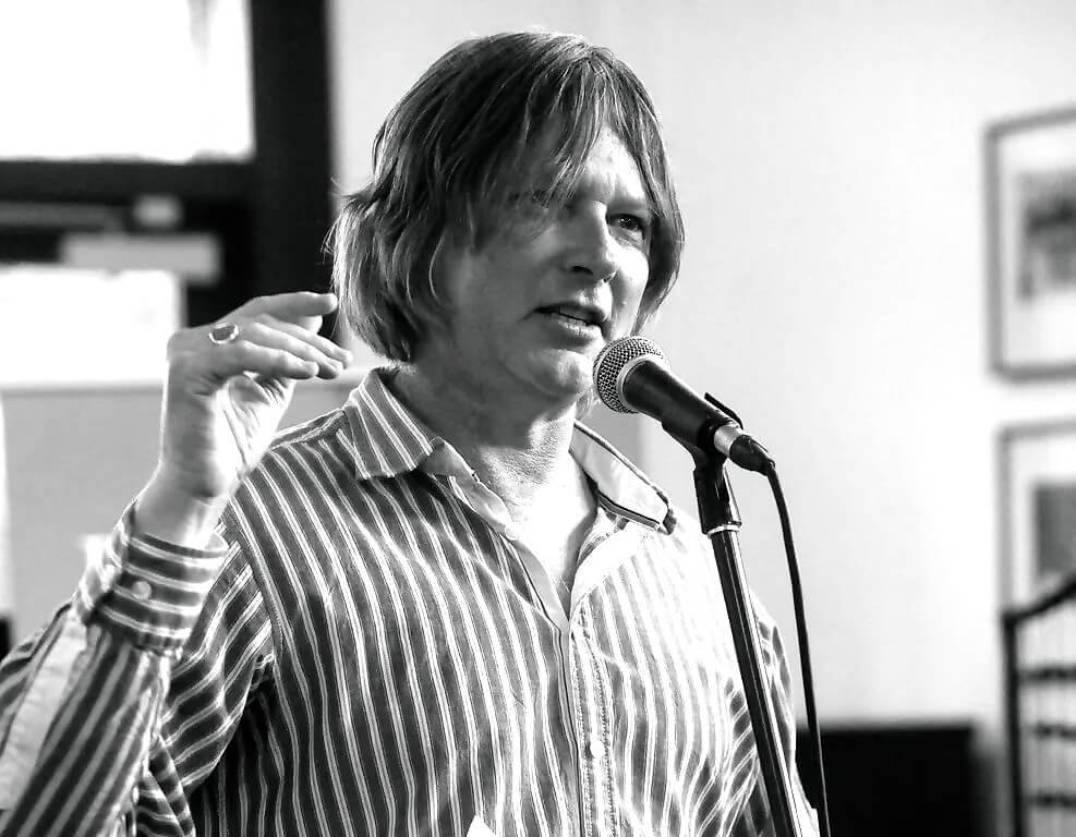 A woman with shoulder-length hair speaking into a microphone during a presentation or speech.