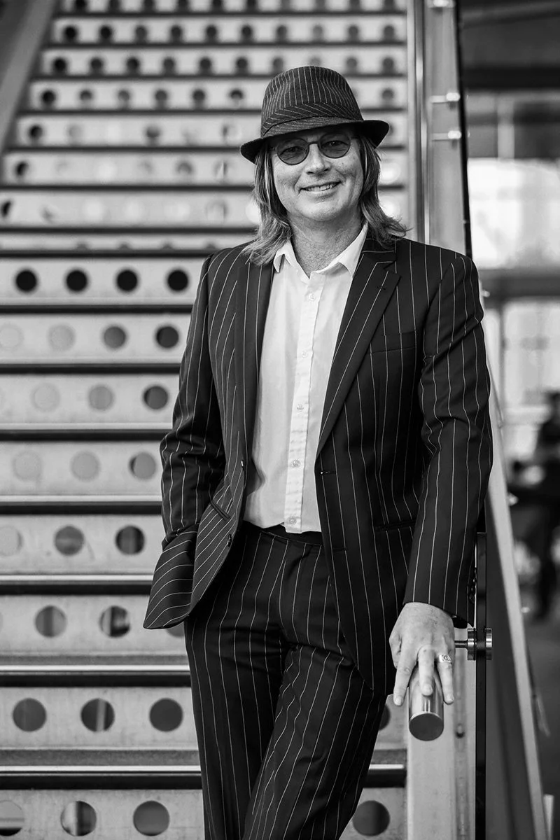 A woman in a pinstripe suit and fedora hat standing on stairs, smiling at the camera in a black and white photo.