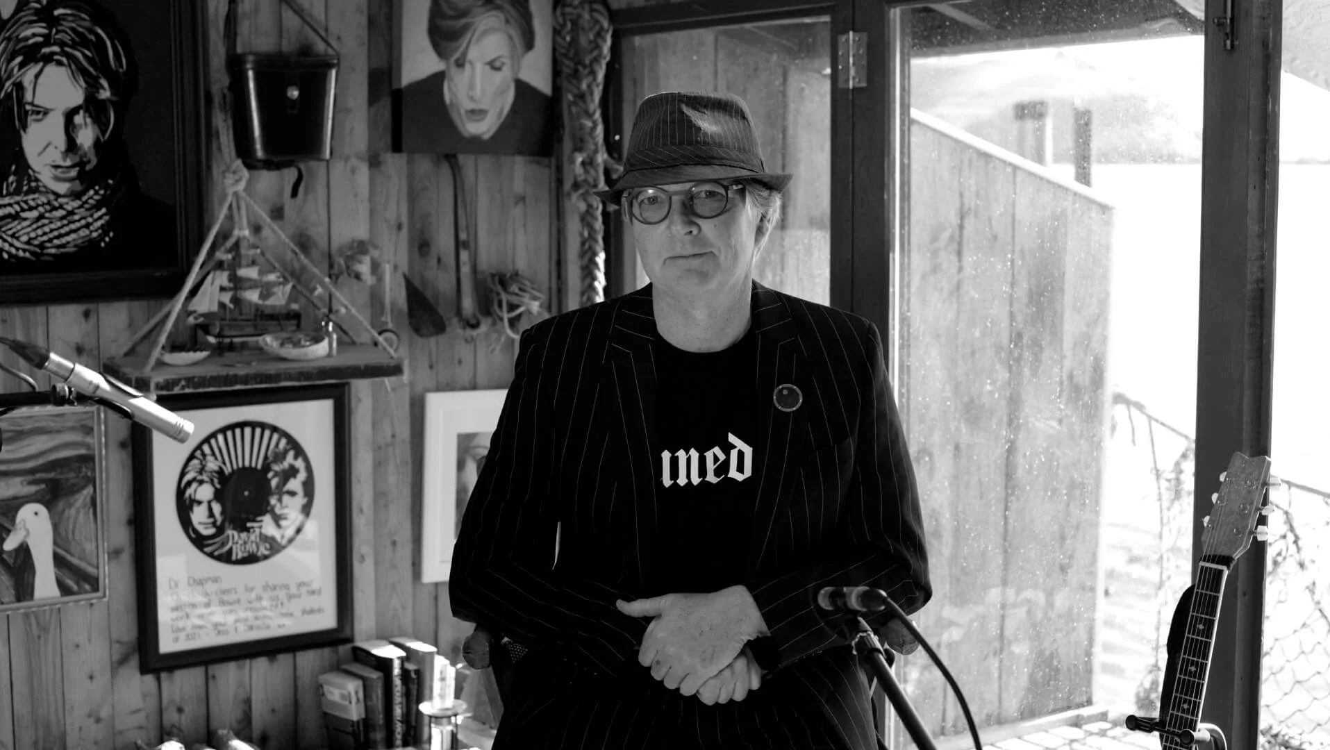 A man wearing glasses, a fedora, and a pinstripe blazer stands in a room with wooden walls, surrounded by artwork and musical instruments, with a window in the background.