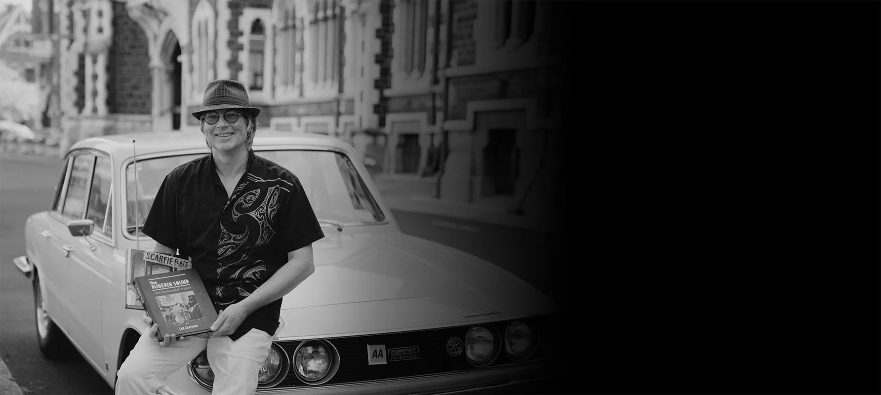 A woman with glasses and a hat leaning against a vintage car on a city street, holding books.