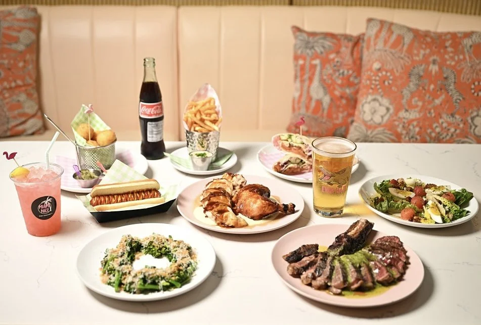 Miami Nice new American restaurant table spread with roasted chicken, juicy ribeye steak with chimichurri, homemade croquetas, kosher hot dog, fries, fresh salad, Mexican Coca-Cola bottle, draft beer, and pink lemonade in Miami Design District.