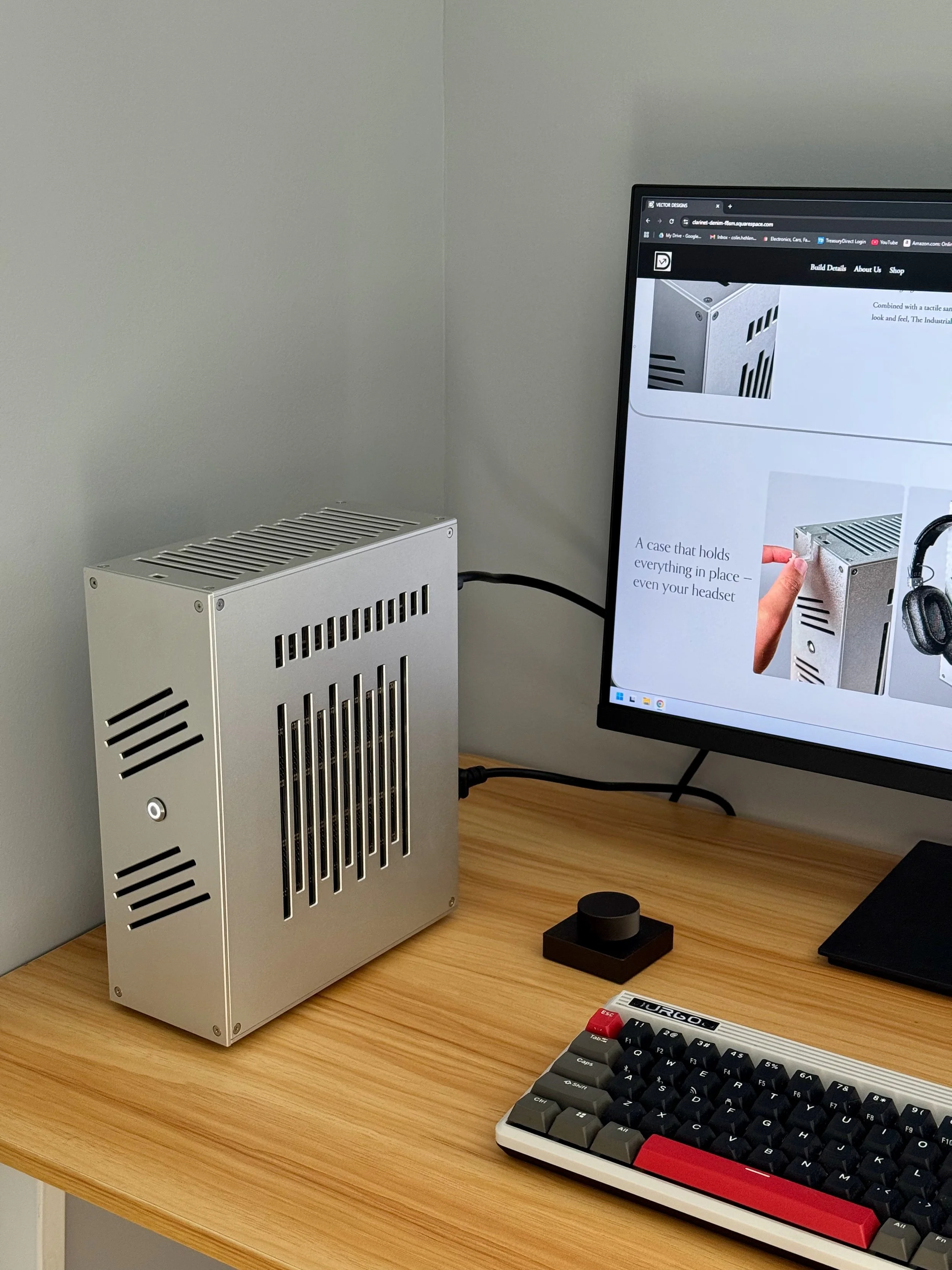 A desk setup featuring a silver computer case, a monitor displaying a webpage with images of computer cases, a black trackball mouse, a vintage keyboard with black, gray, and red keys, a black mouse pad, and a small black speaker on a wooden desk aga