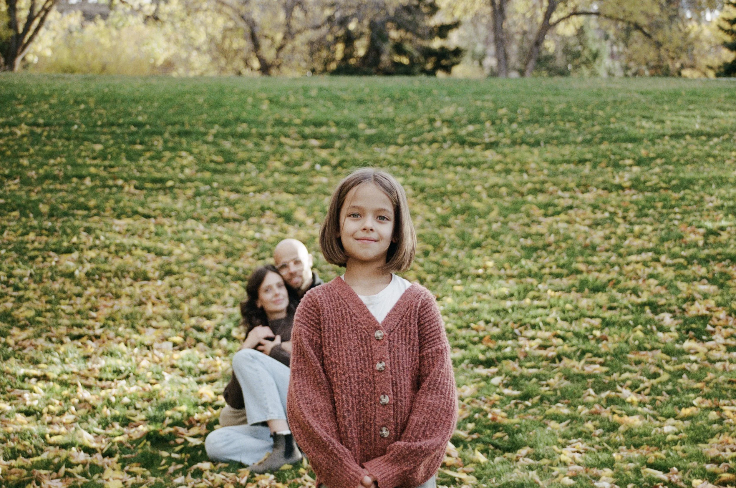A young girl with shoulder-length brown hair standing in front of a family of three sitting on the grass in a park during autumn. The girl is smiling and wearing a red cardigan over a white shirt. The family behind her appears to be a man and a woman