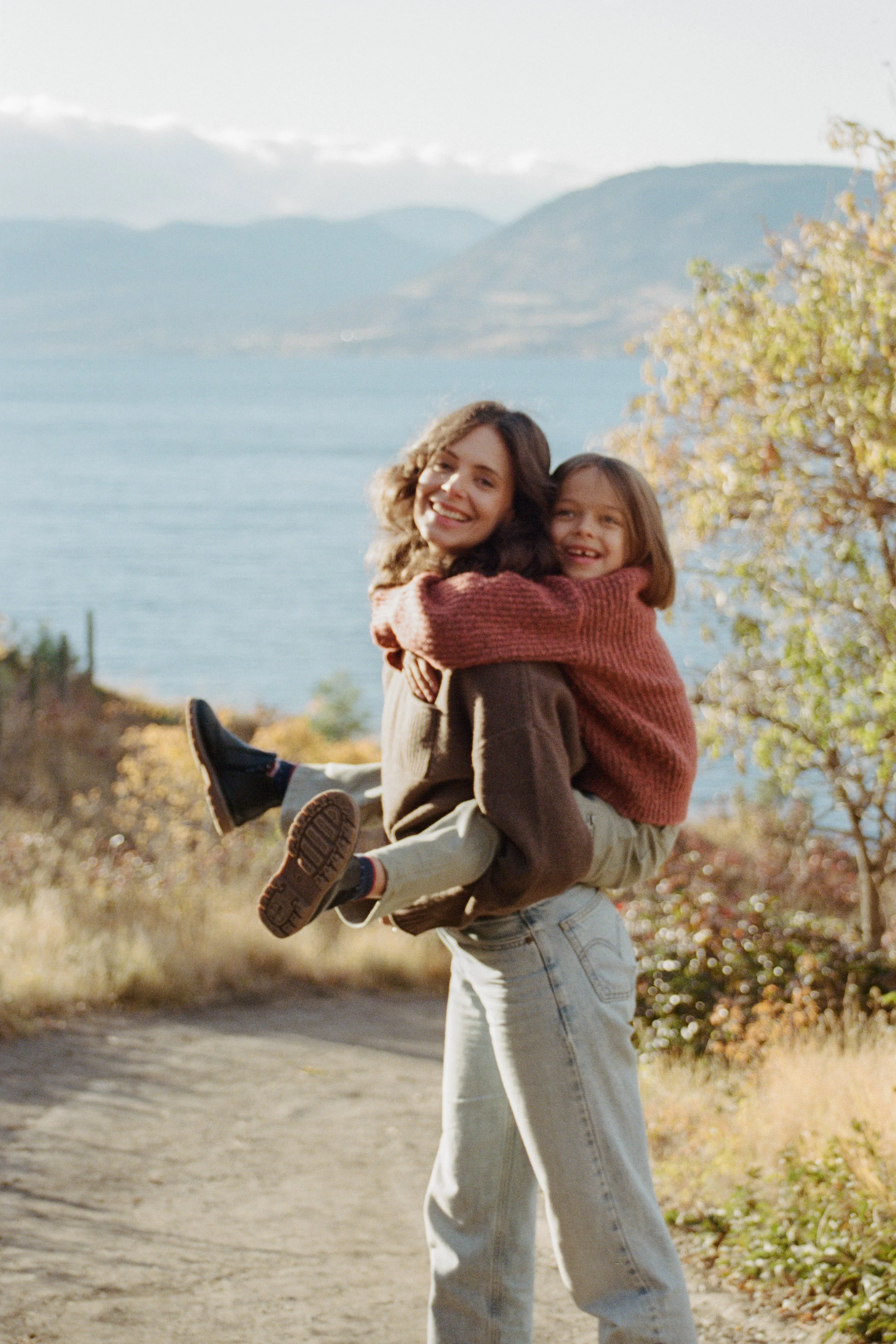 A woman holding a young girl in her arms outdoors during autumn, with a lake, mountains, and trees in the background.