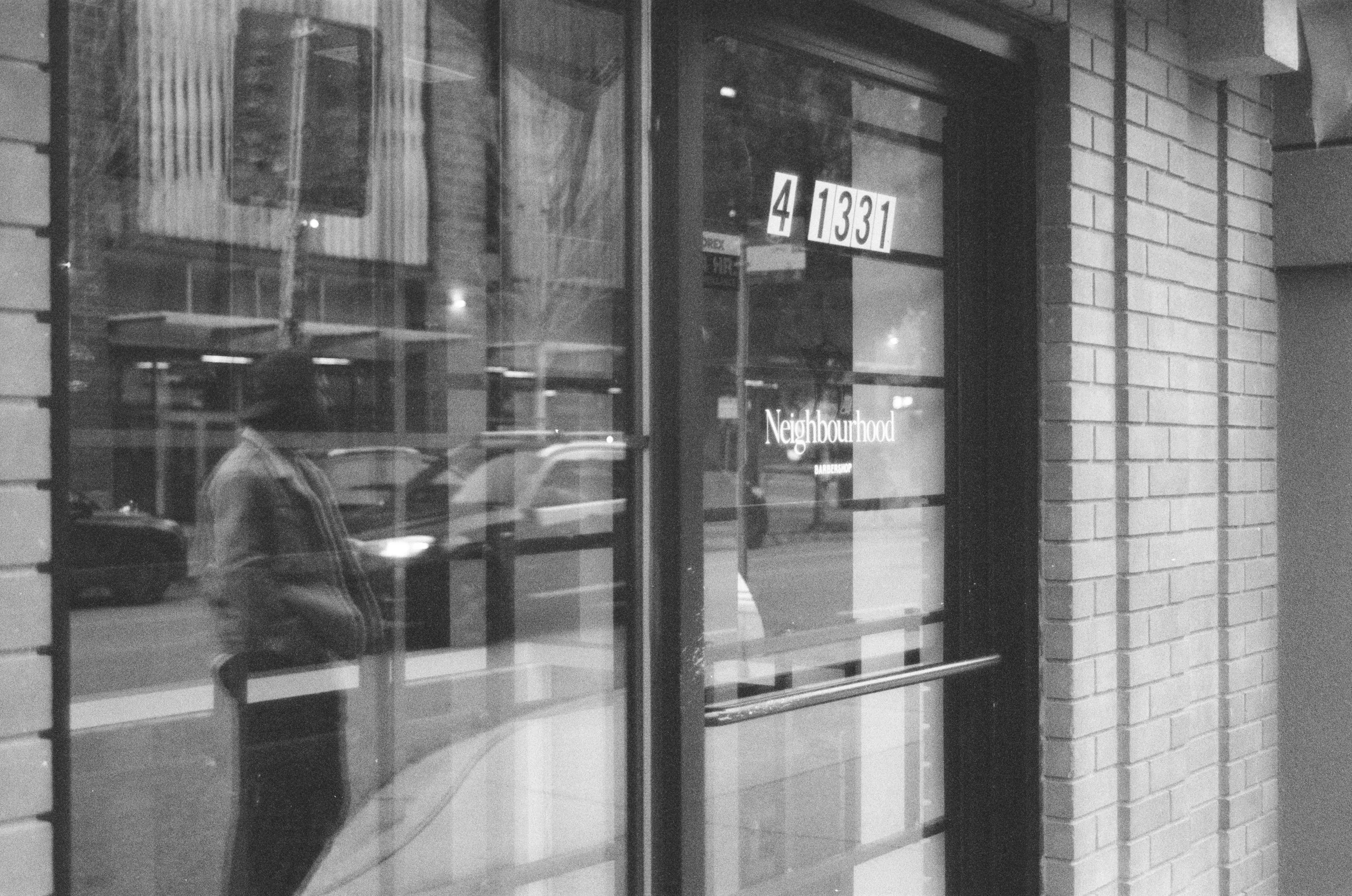 Black and white photo of a storefront window with the address "41331" and the word "Neighbourhood" on the glass. Reflections of a person looking at their phone and trees are visible in the window.