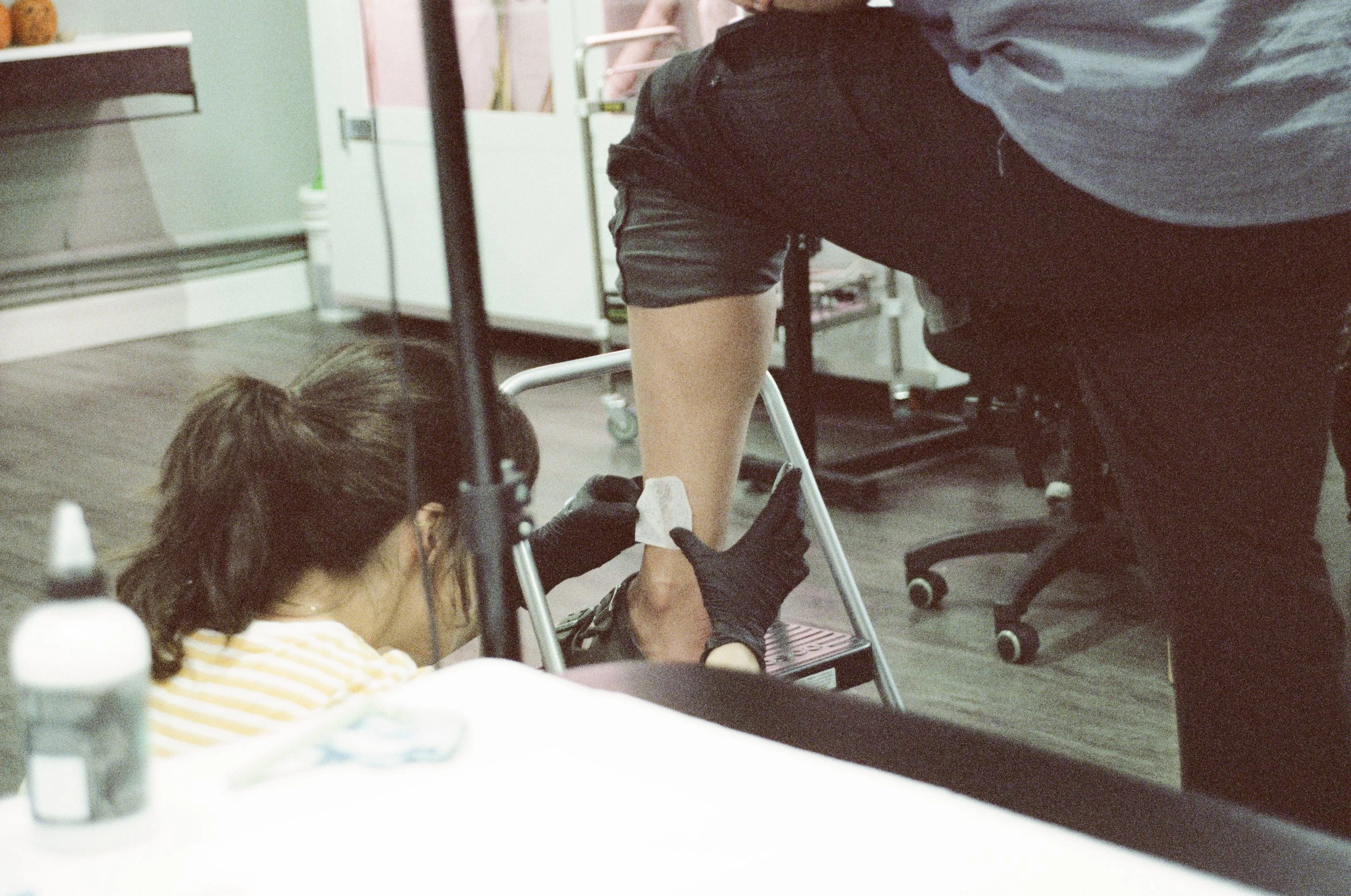 A person receiving a tattoo on their arm from a tattoo artist in a studio. The artist is wearing black gloves and working on the person's forearm, which is resting on a small stool. Various tattoo supplies are visible in the foreground.