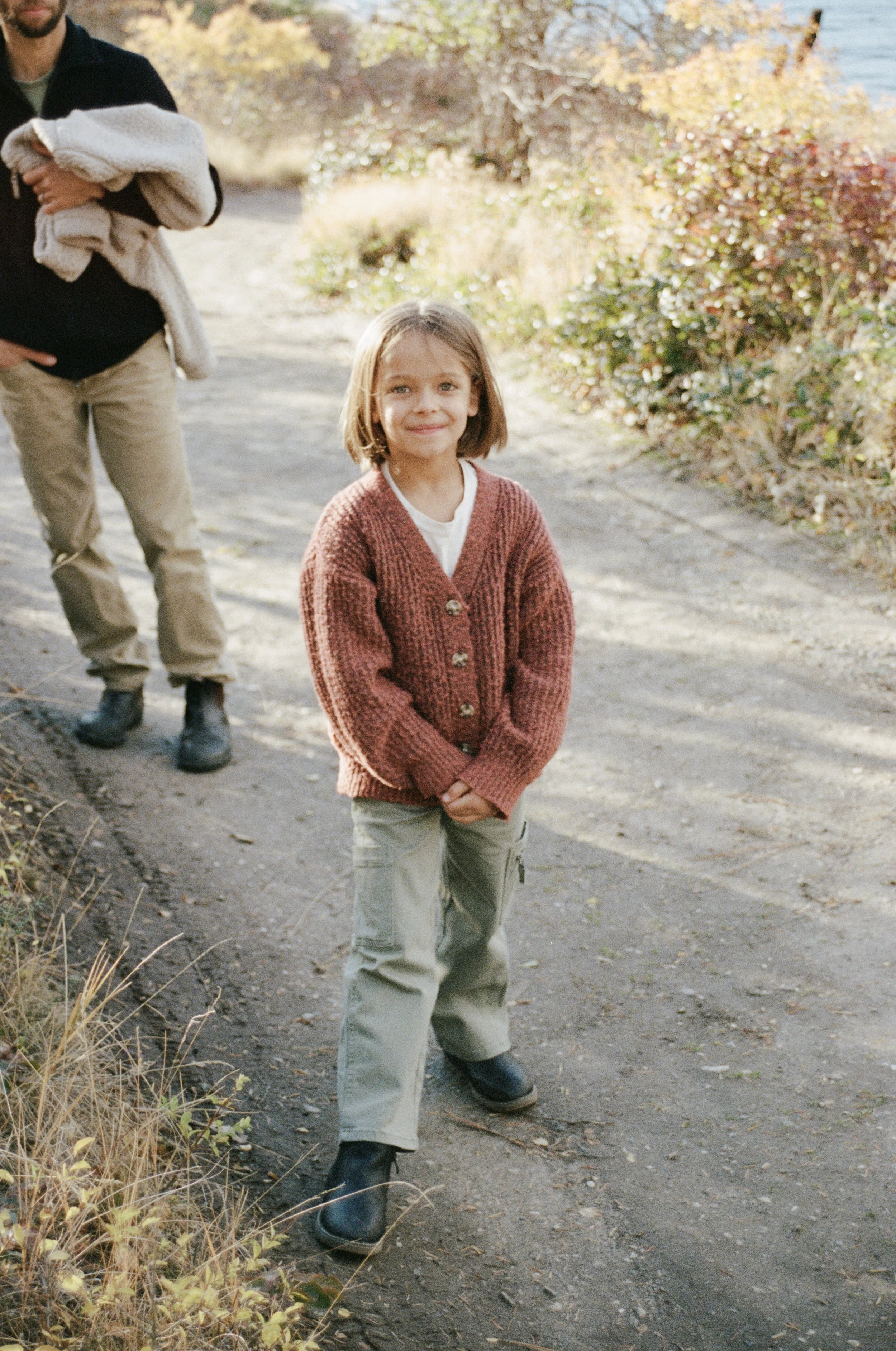 A young girl standing on a dirt path outdoors with autumn foliage in the background, smiling at the camera, wearing a red cardigan, white shirt, beige cargo pants, and black boots. A man stands nearby holding a blanket.